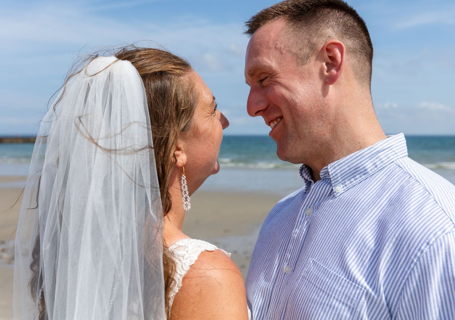 A bride and groom share a joyful moment together on a sunny beach with ocean waves in the background.