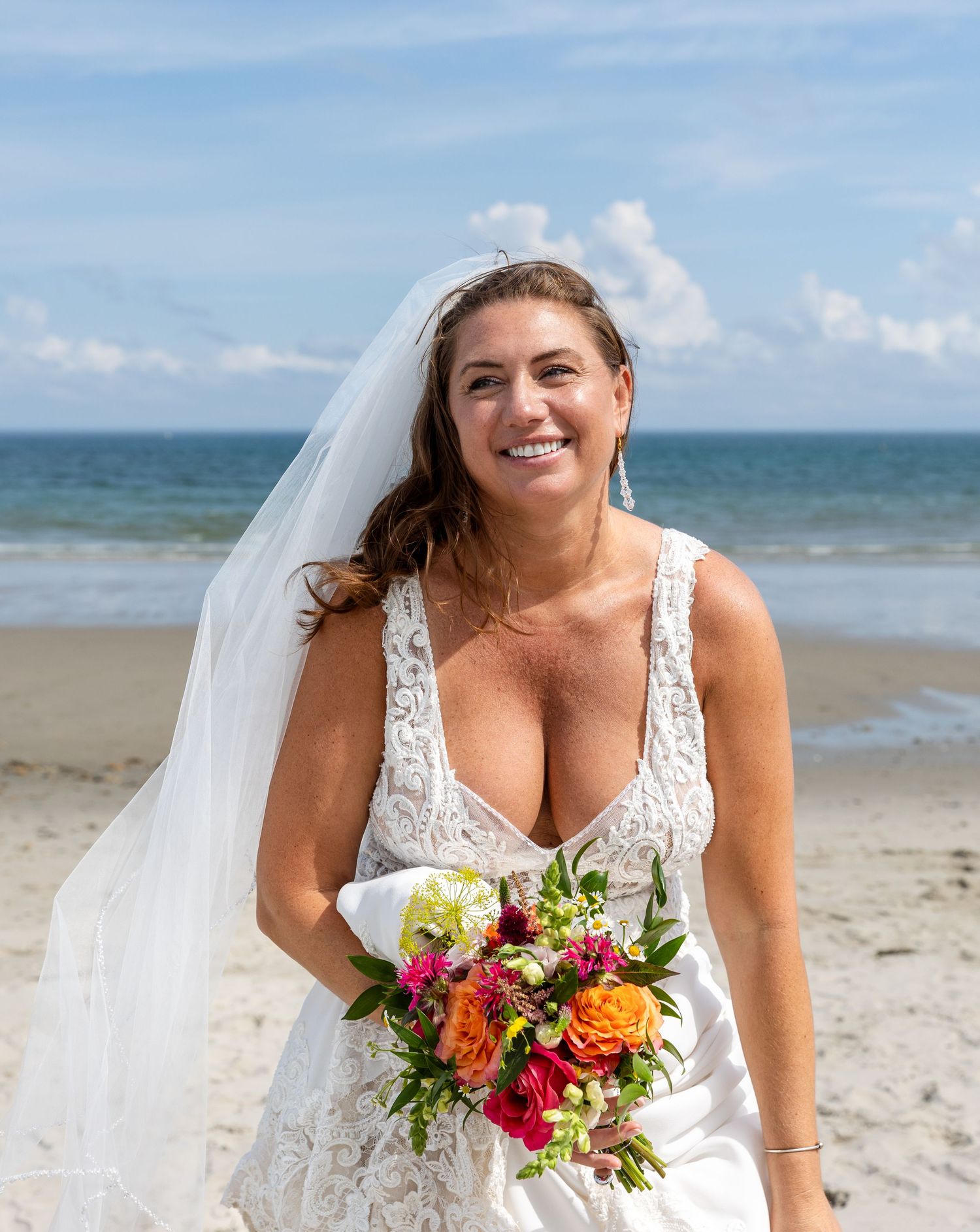 A bride in a lace wedding dress holds a vibrant bouquet of orange, red and pink flowers on a sunny beach.