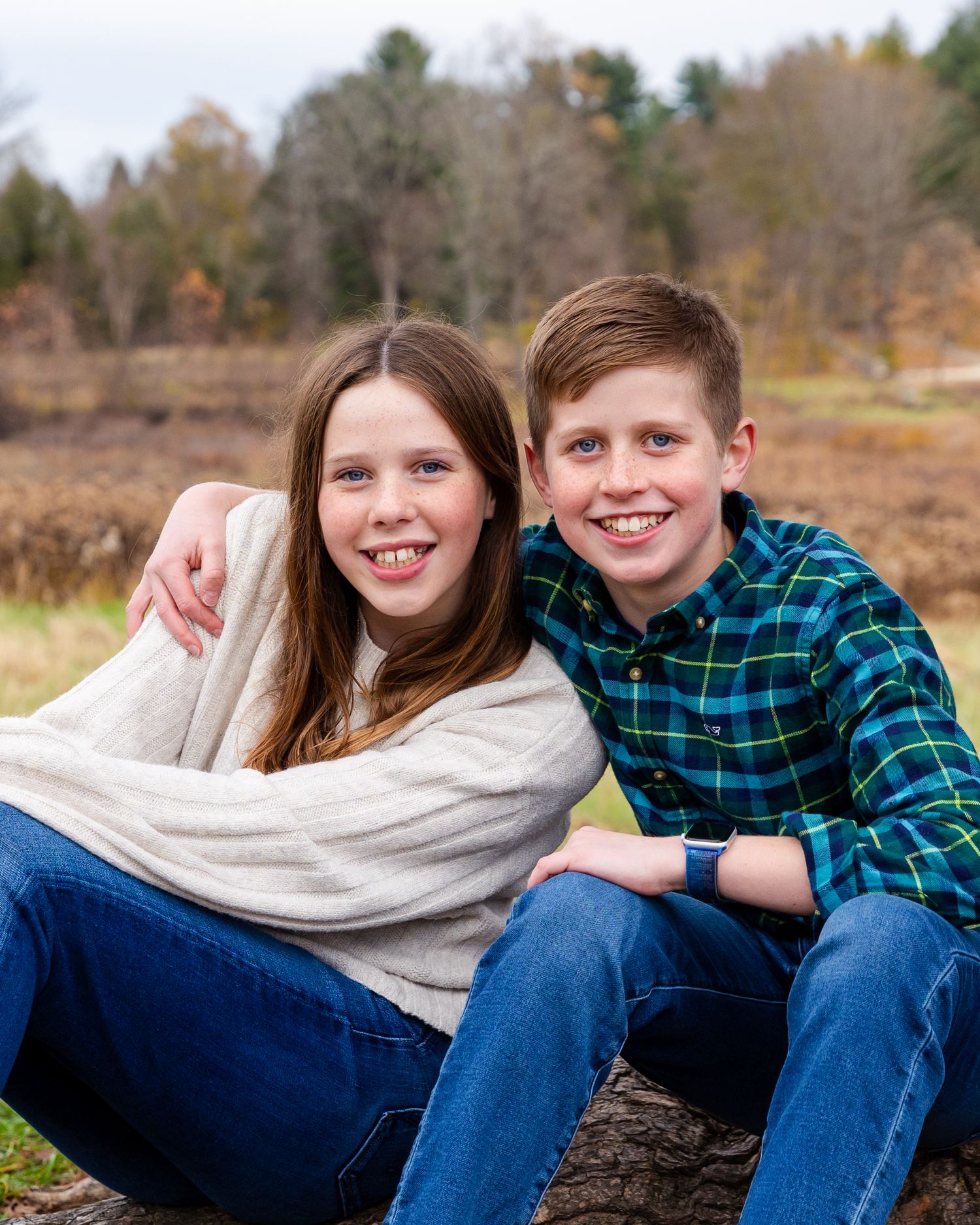 Young siblings in jeans sit together outdoors on fallen log with autumn trees and field in background.