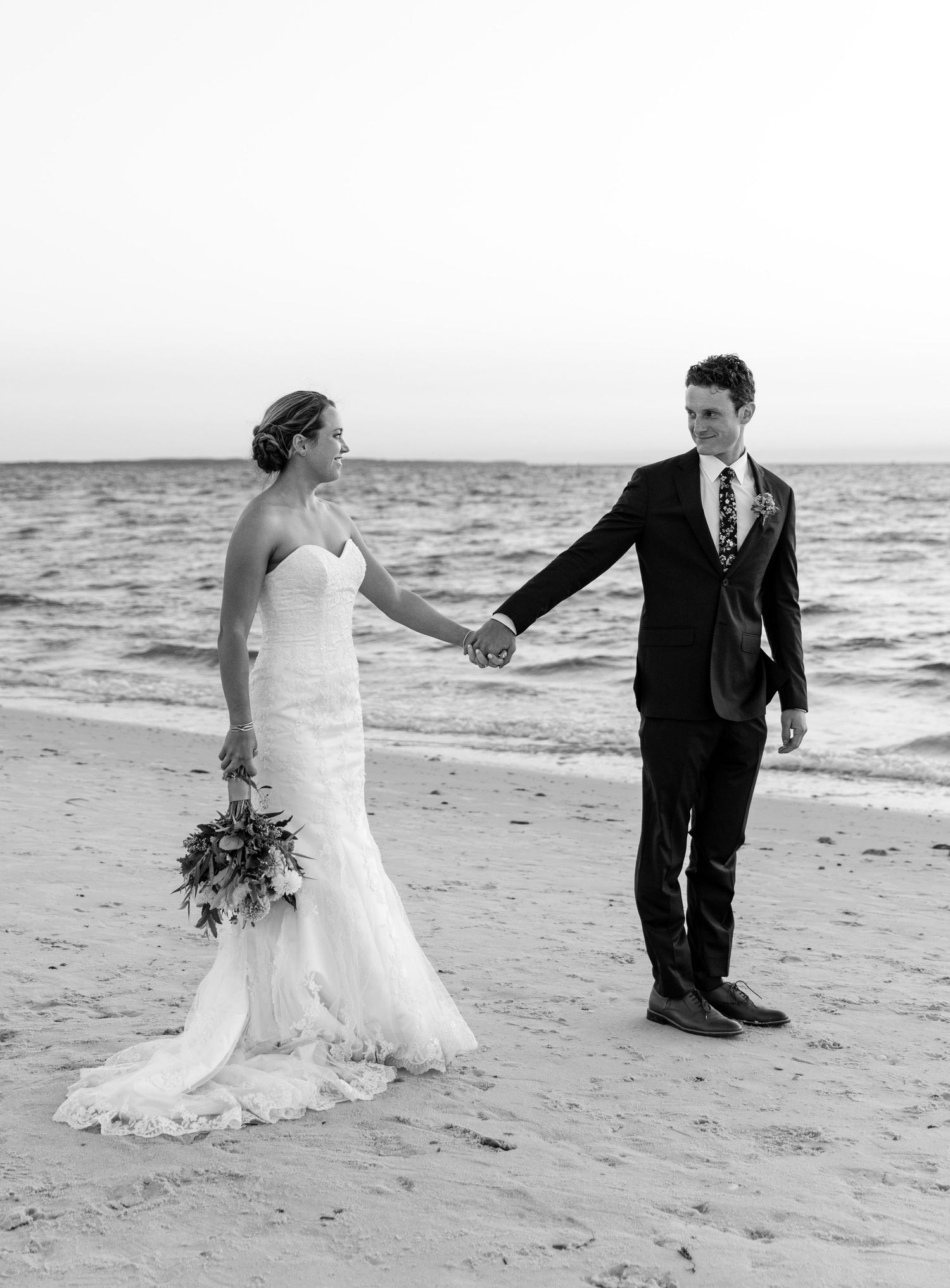 Black and white photos of newlyweds walking together on a sandy beach with ocean waves in the background.