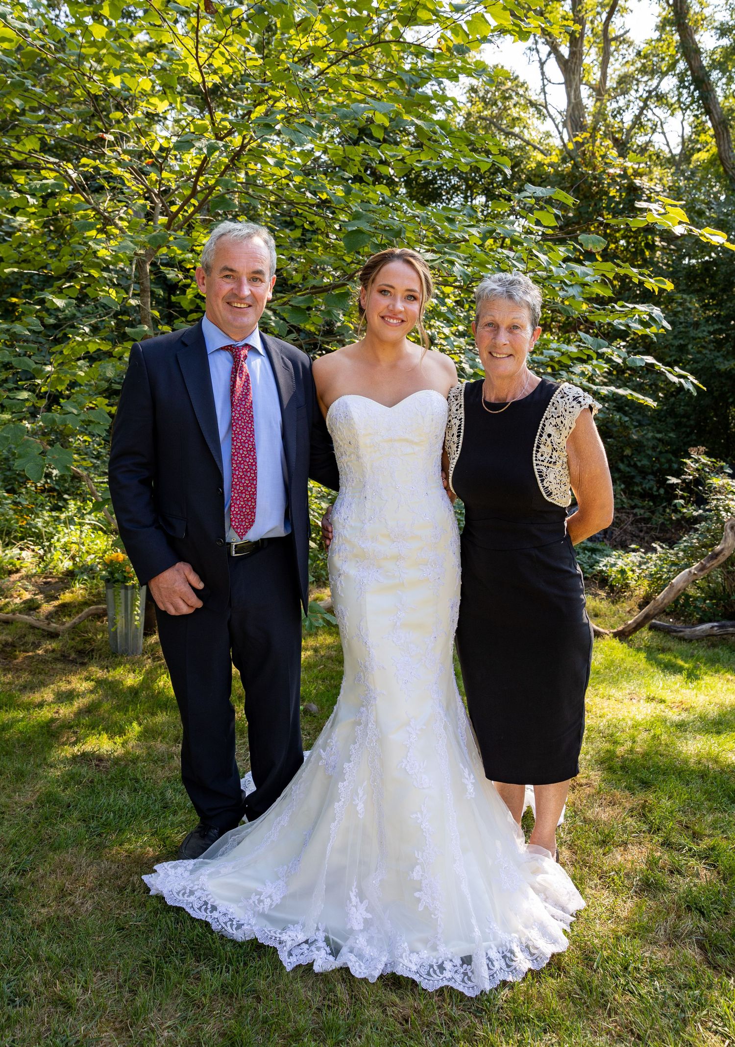 A wedding party poses outdoors against lush greenery with the bride wearing an elegant mermaid style gown.