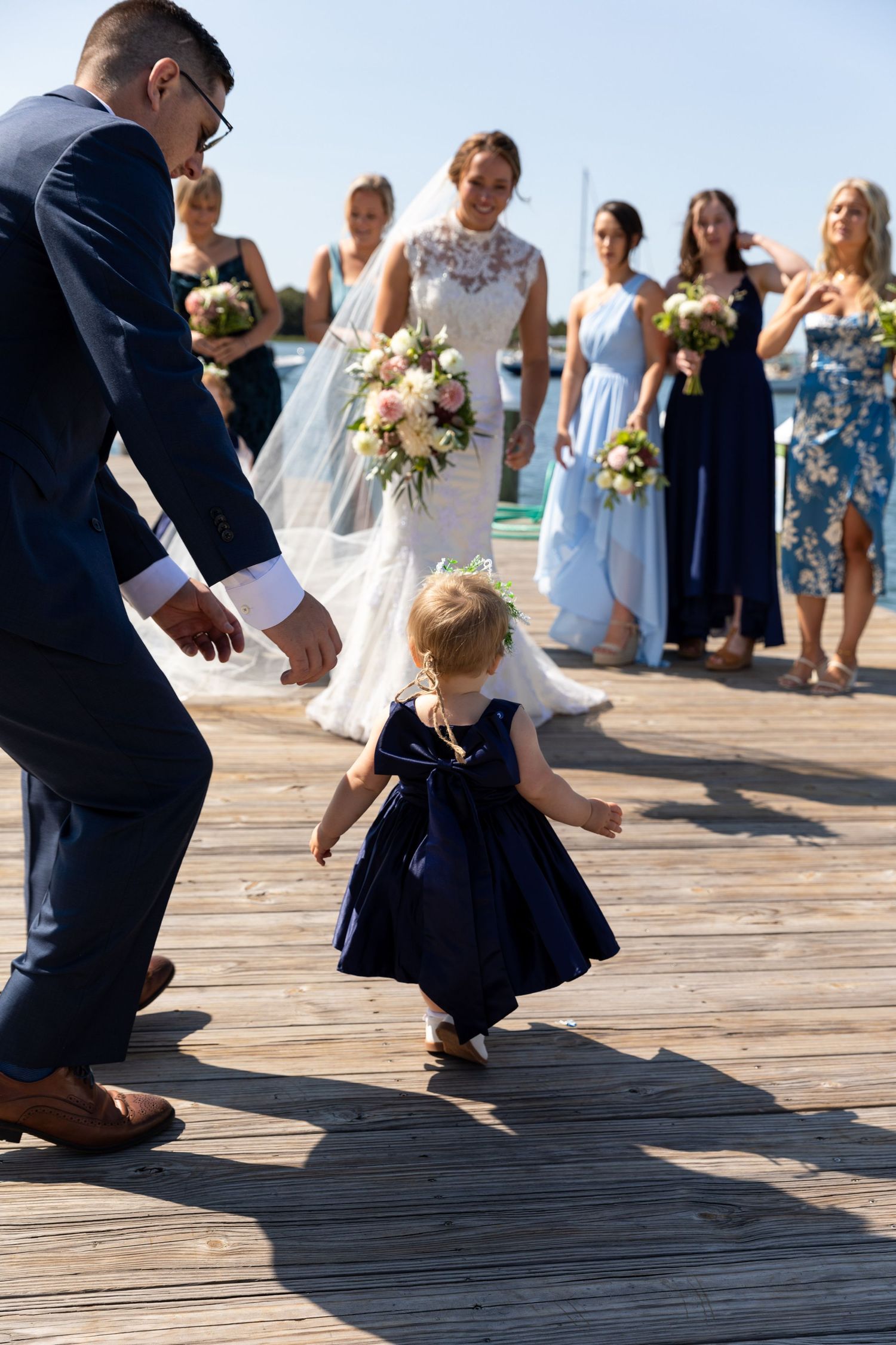 A small child in a navy dress dances on a wooden deck during a wedding celebration with guests watching.