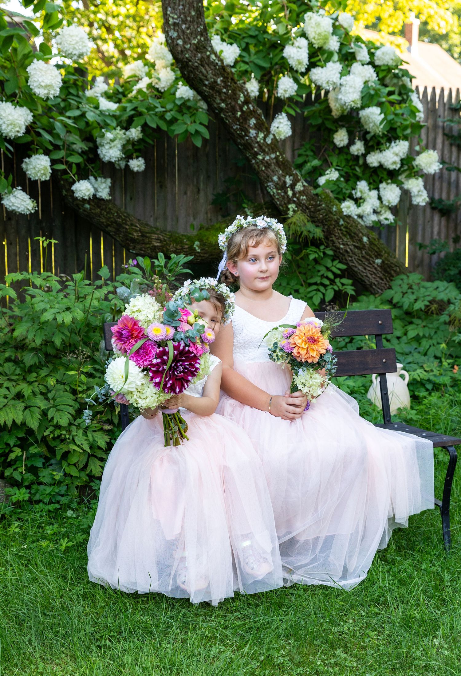 Two flower girls in white tulle dresses sit on a garden bench holding vibrant floral bouquets under blooming white roses.