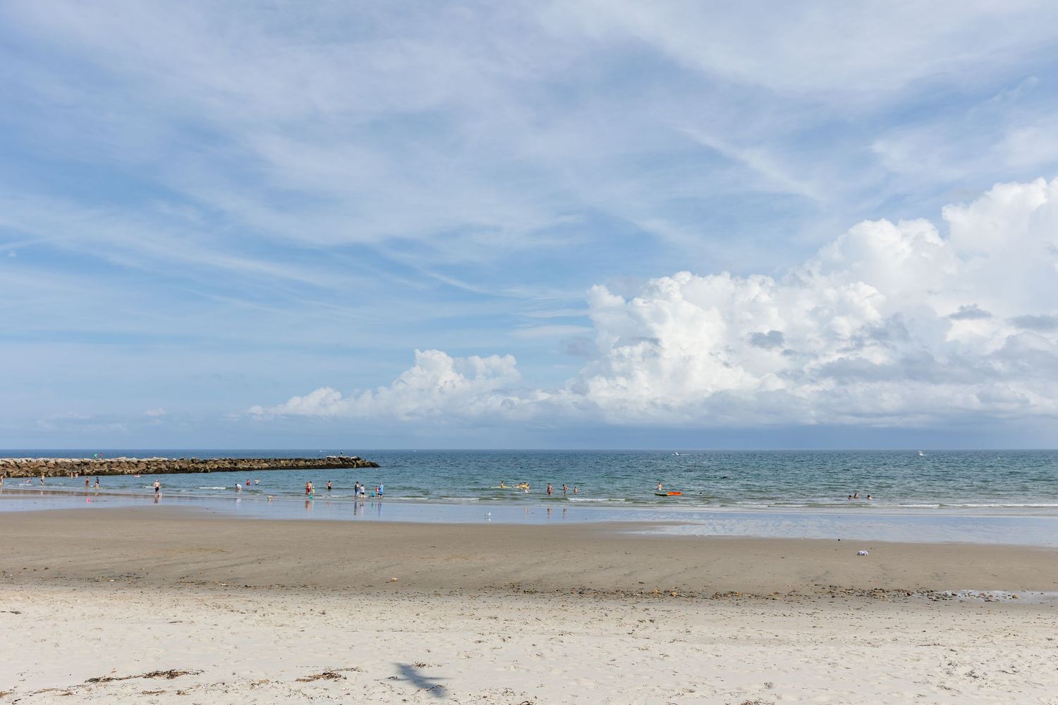 Panoramic view of a sandy beach with a long wooden pier extending into the ocean under cloudy blue skies.