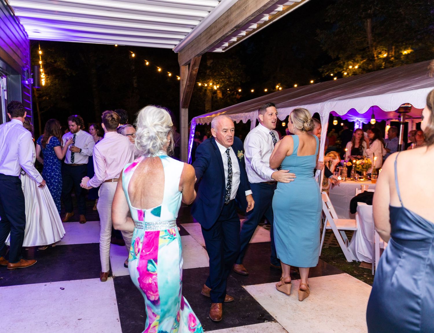 Wedding guests dancing and mingling on an illuminated outdoor patio with purple lighting and string lights overhead.