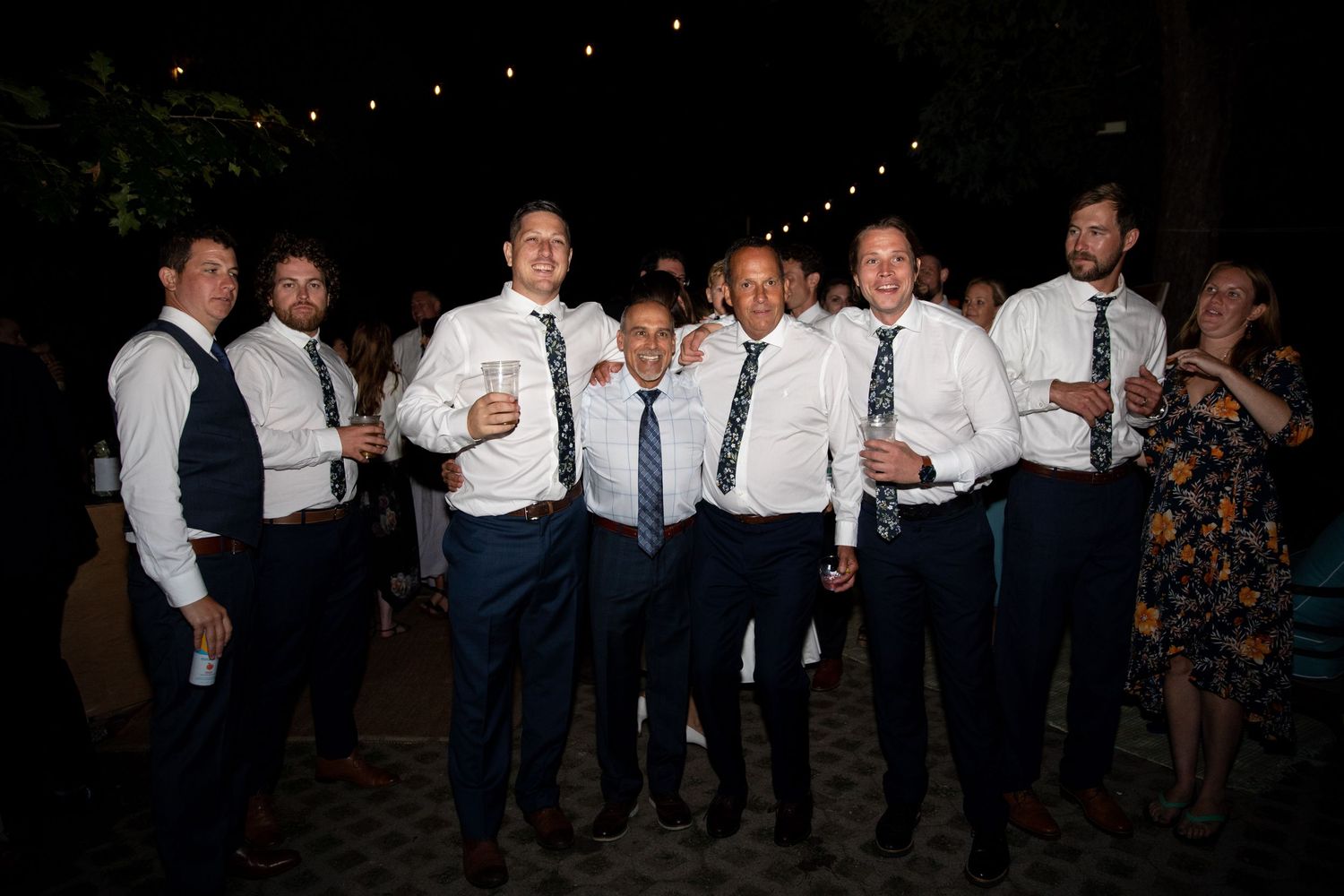 A group of groomsmen in white shirts and navy ties gather together for a nighttime outdoor celebration.