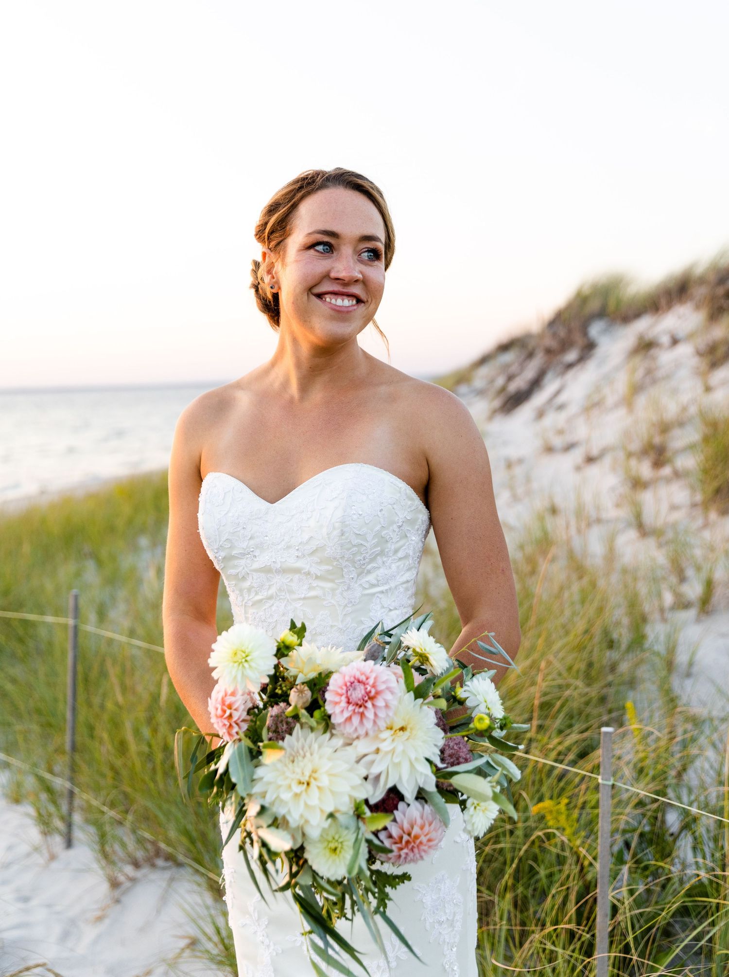 A gorgeous wedding bouquet with pink and white flowers held against a sandy beach dune background at sunset.