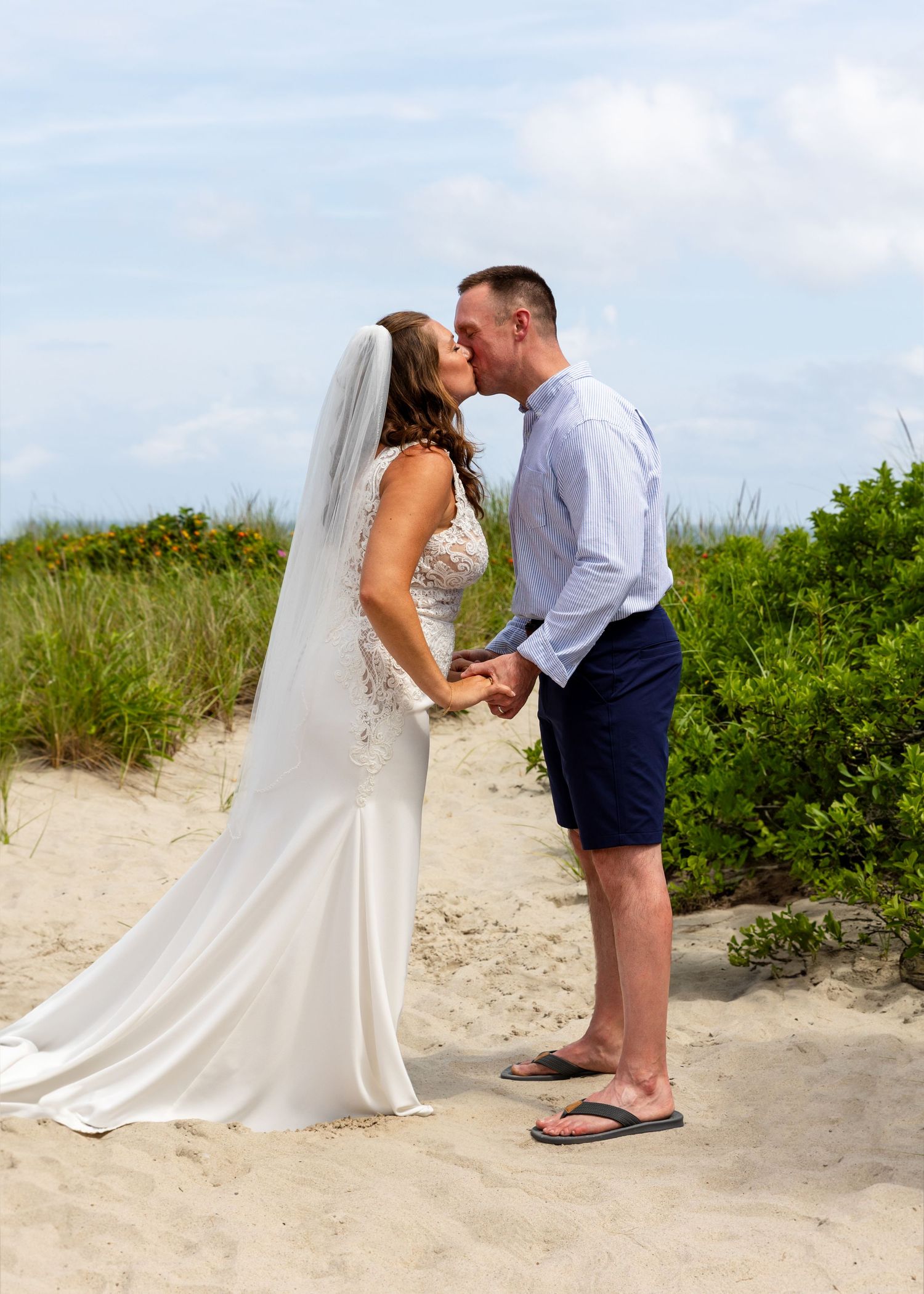 A couple shares a romantic kiss on a scenic beach path with coastal vegetation and sandy dunes in the background.