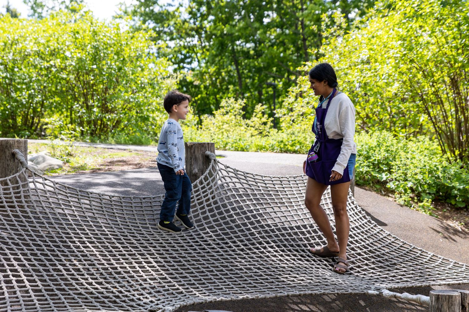 Two people playing together on a large outdoor rope net installation surrounded by green foliage.