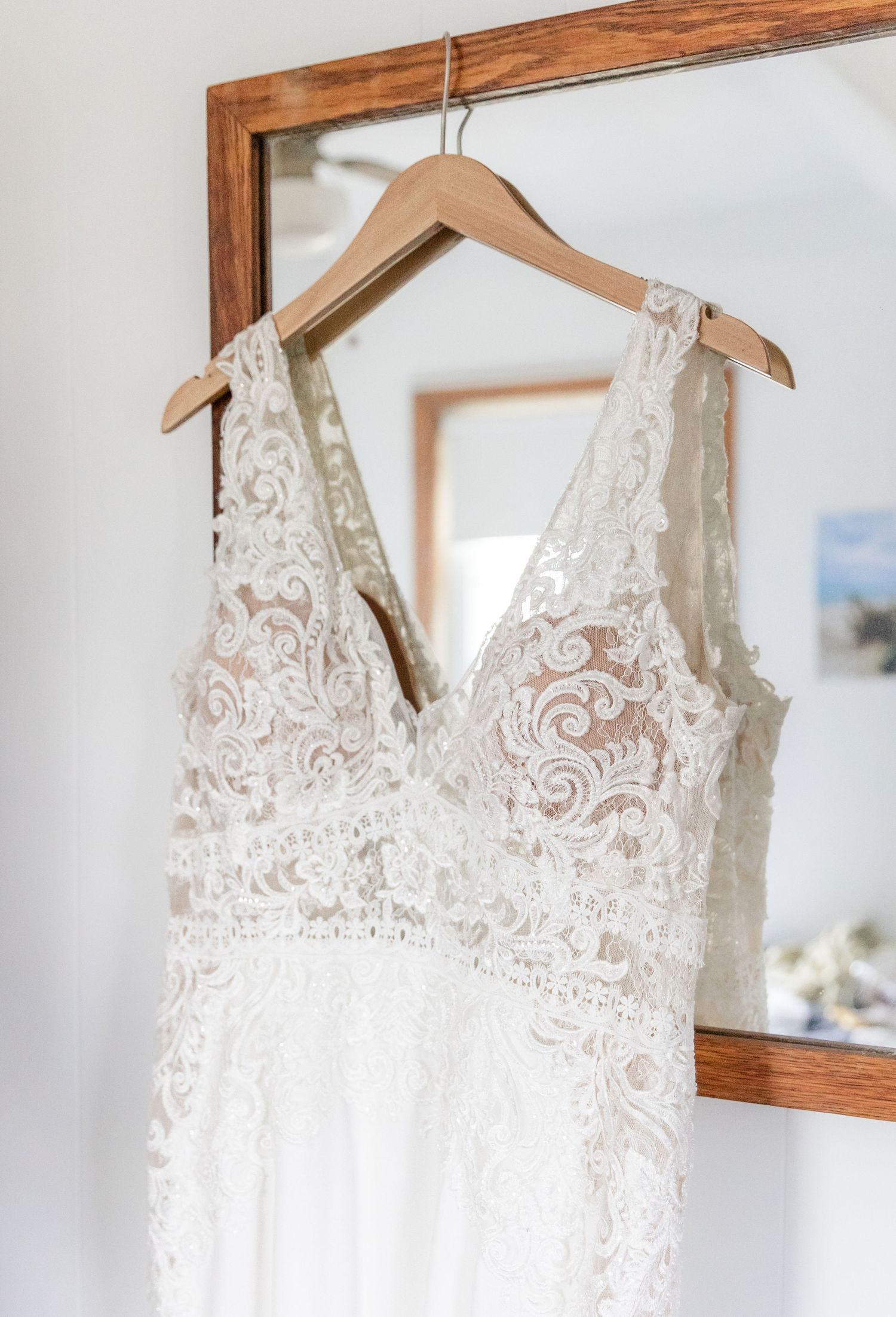 White lace wedding dress with intricate floral pattern and deep v-neckline hanging on wooden hanger against wooden frame.