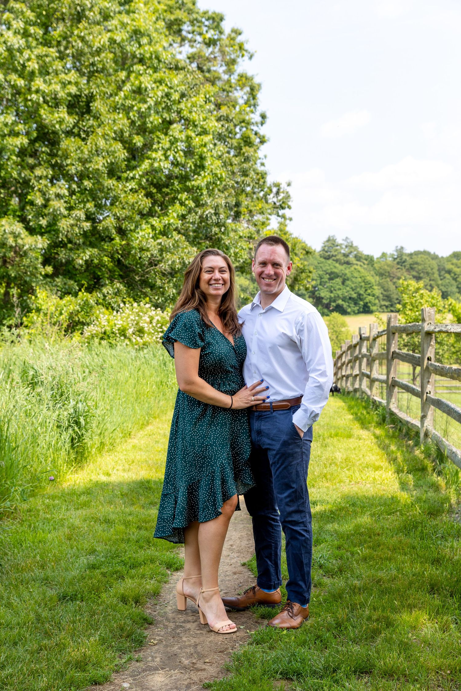 A couple enjoys a romantic outdoor photoshoot along a rustic wooden fence on a sunny summer day with lush greenery.
