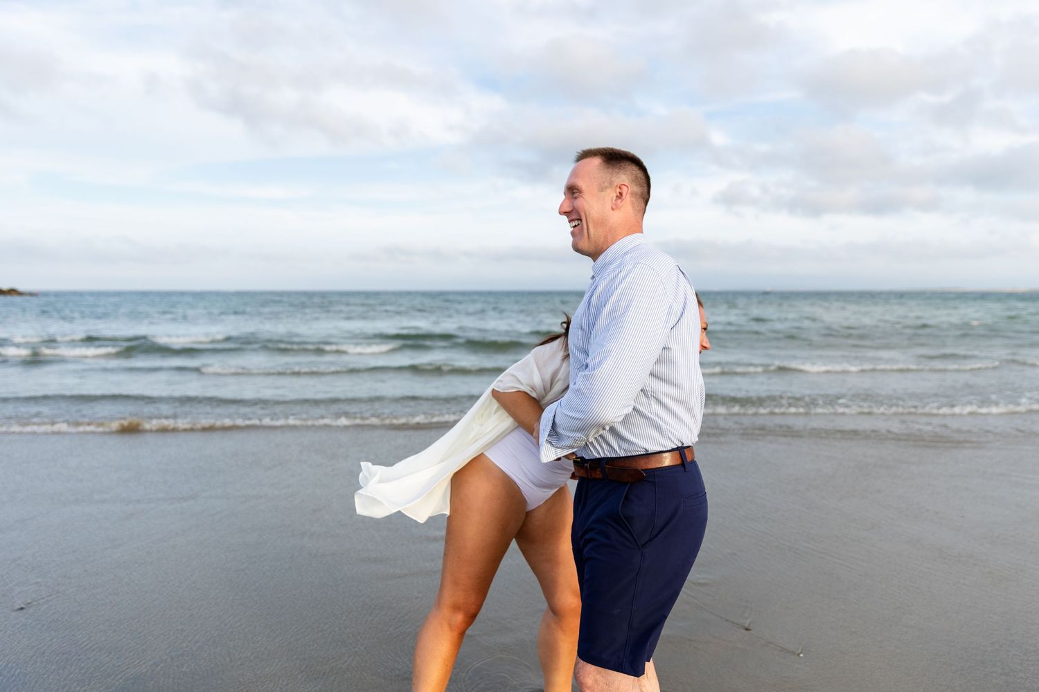 A couple embraces playfully on a cloudy beach while standing near the ocean waves.