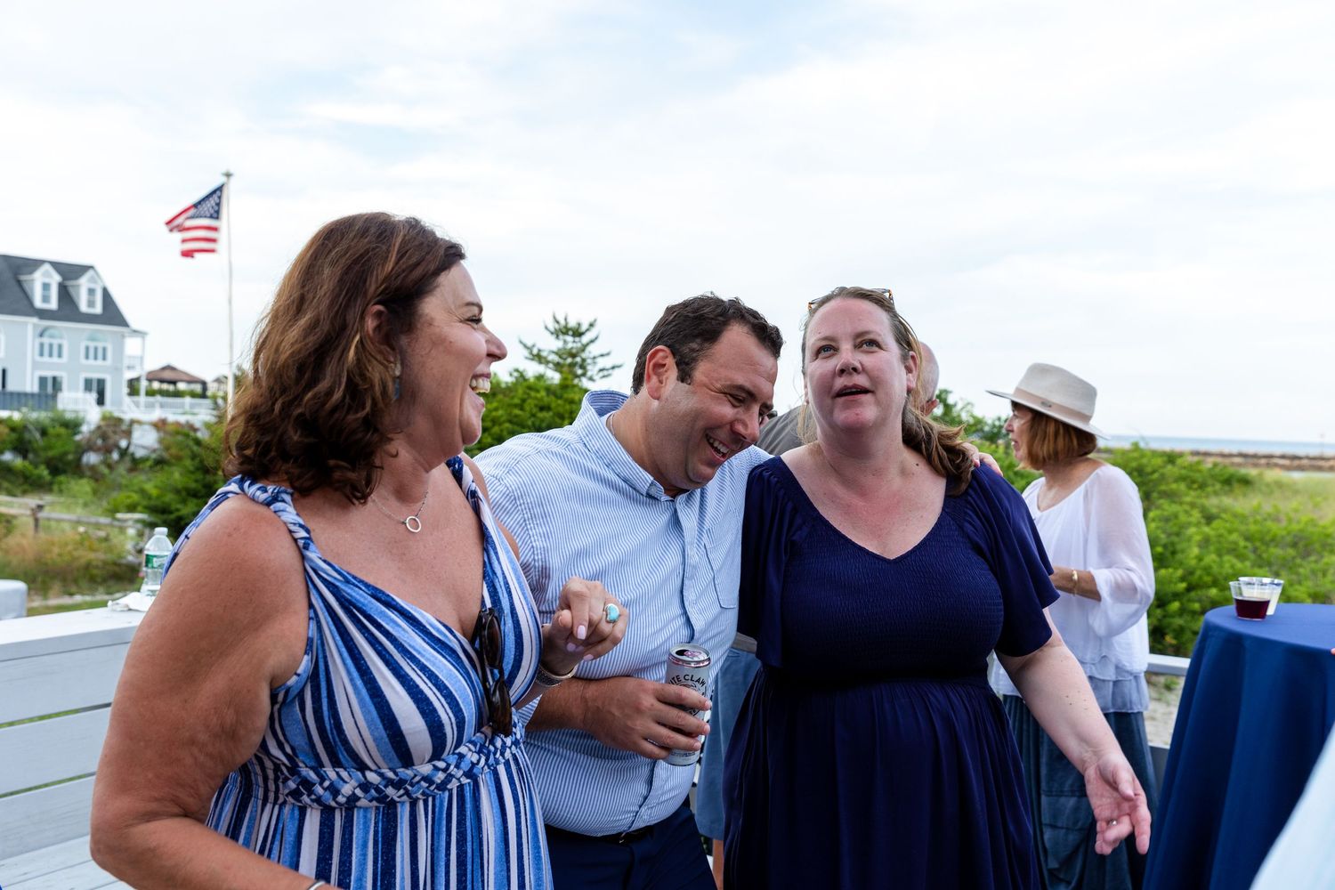 Three people converse and laugh together at an outdoor gathering with a flag and coastal scenery in the background.