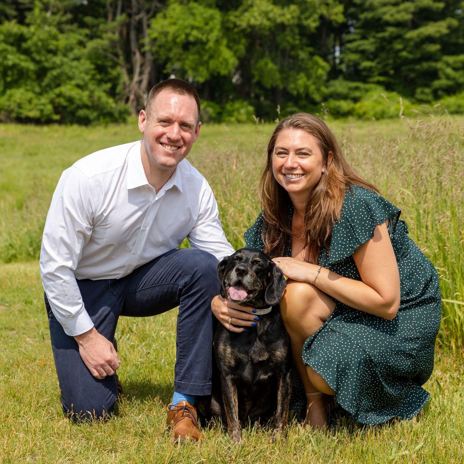 A couple poses in a grassy field with their black Labrador retriever dog during an outdoor photo session.