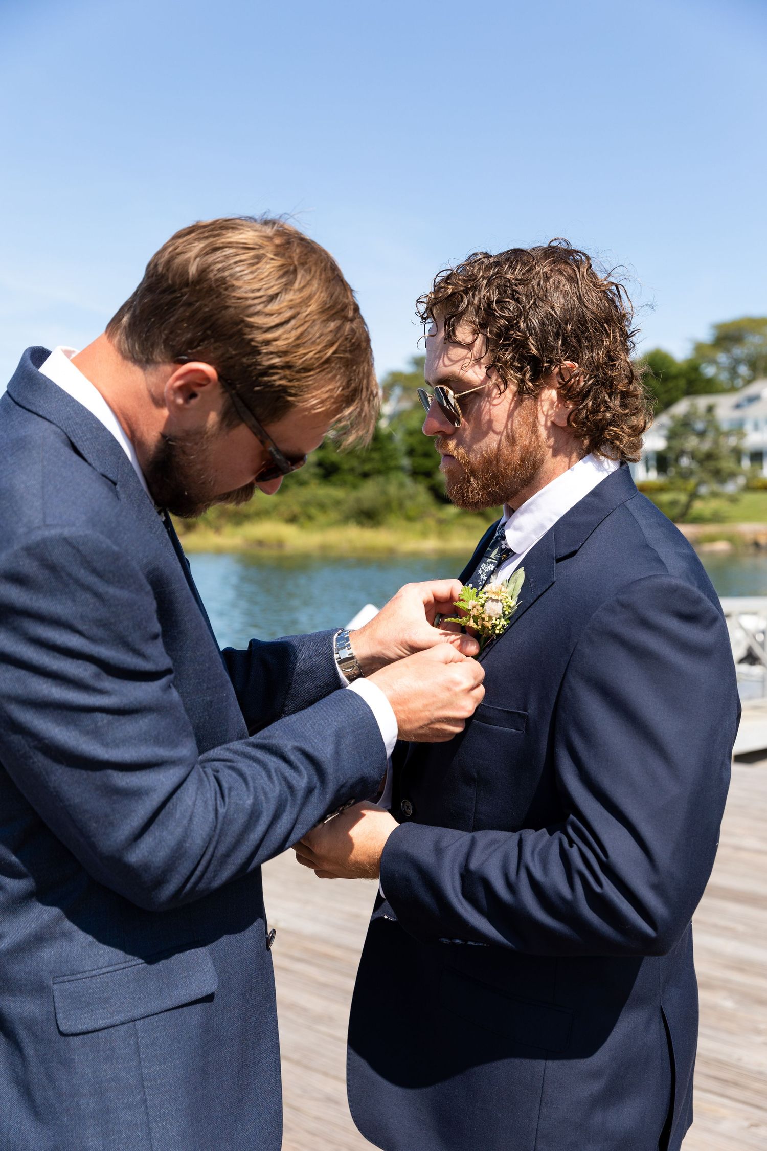 Two people in navy suits helping each other with boutonnieres on a sunny day by the water.