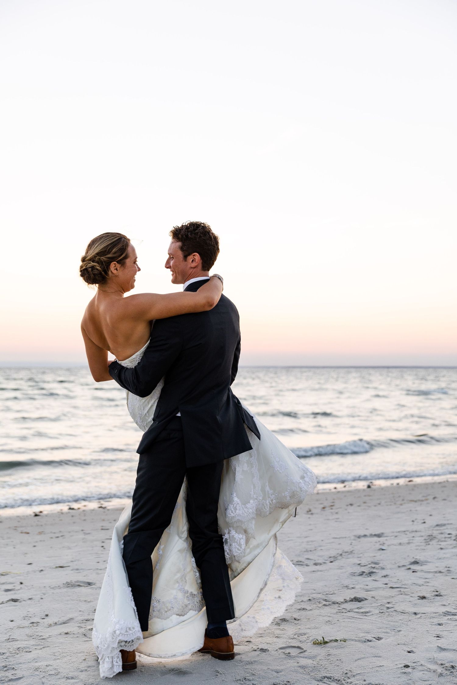 A romantic couple in wedding attire embraces during sunset on a serene beach, walking along the shoreline.