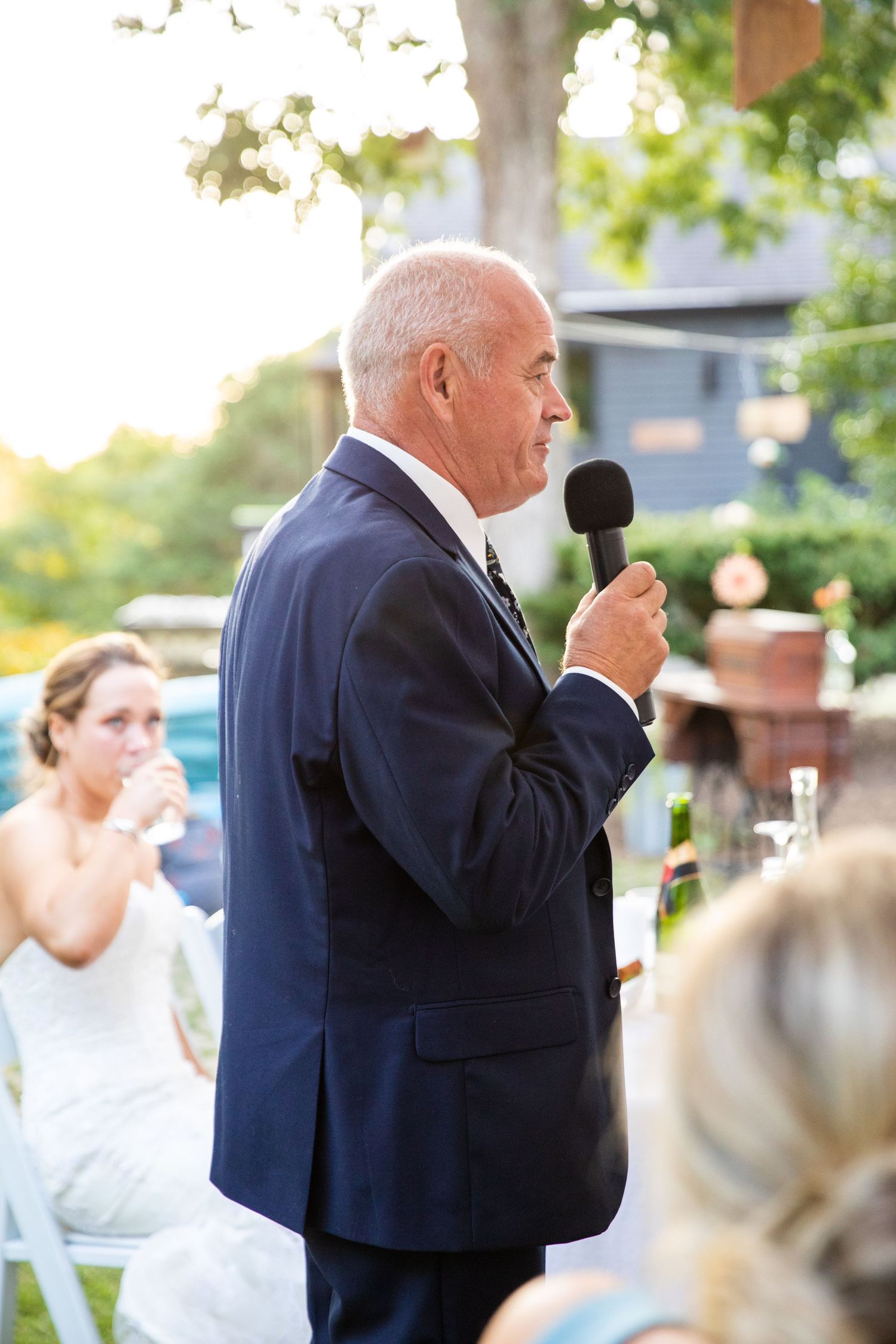 A gentleman in a navy blue suit gives a speech with microphone at an outdoor event with people seated in the background.