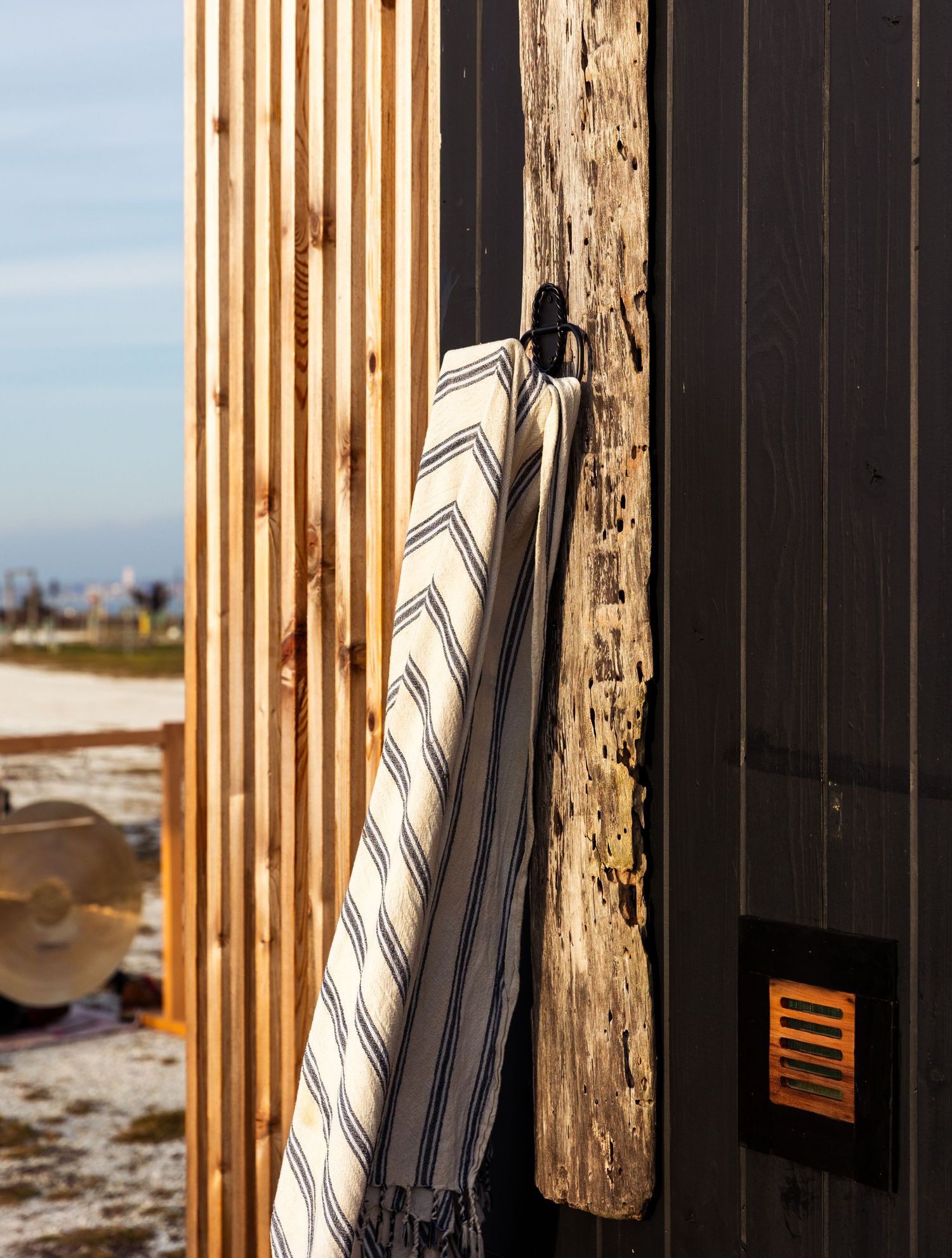 A black and white striped beach towel hangs against a weathered wooden post with industrial buildings in the background.