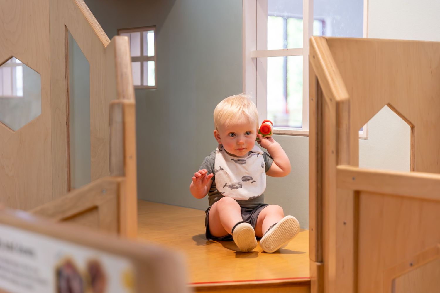 A wooden dollhouse surrounded by various toys with small rooms and windows creates a warm, playful environment.