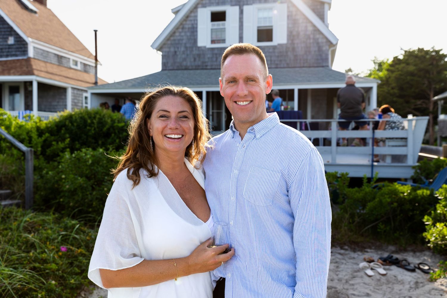 A happy couple stands smiling outdoors in front of a coastal home with white fencing during summer.