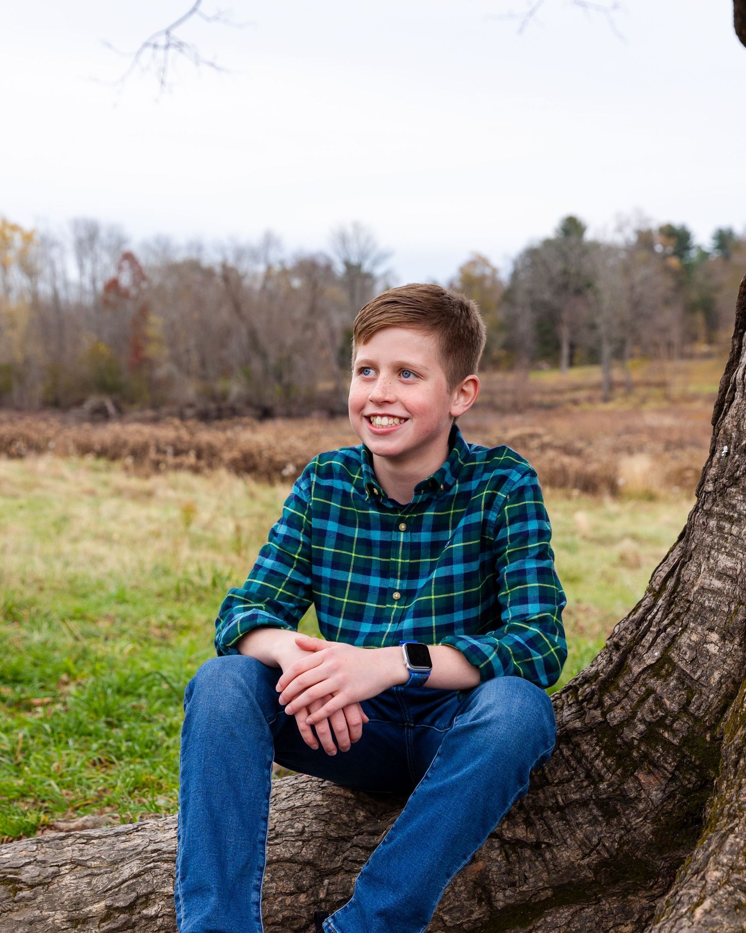 A person in a plaid shirt sits on a curved tree trunk in a natural field setting during autumn.