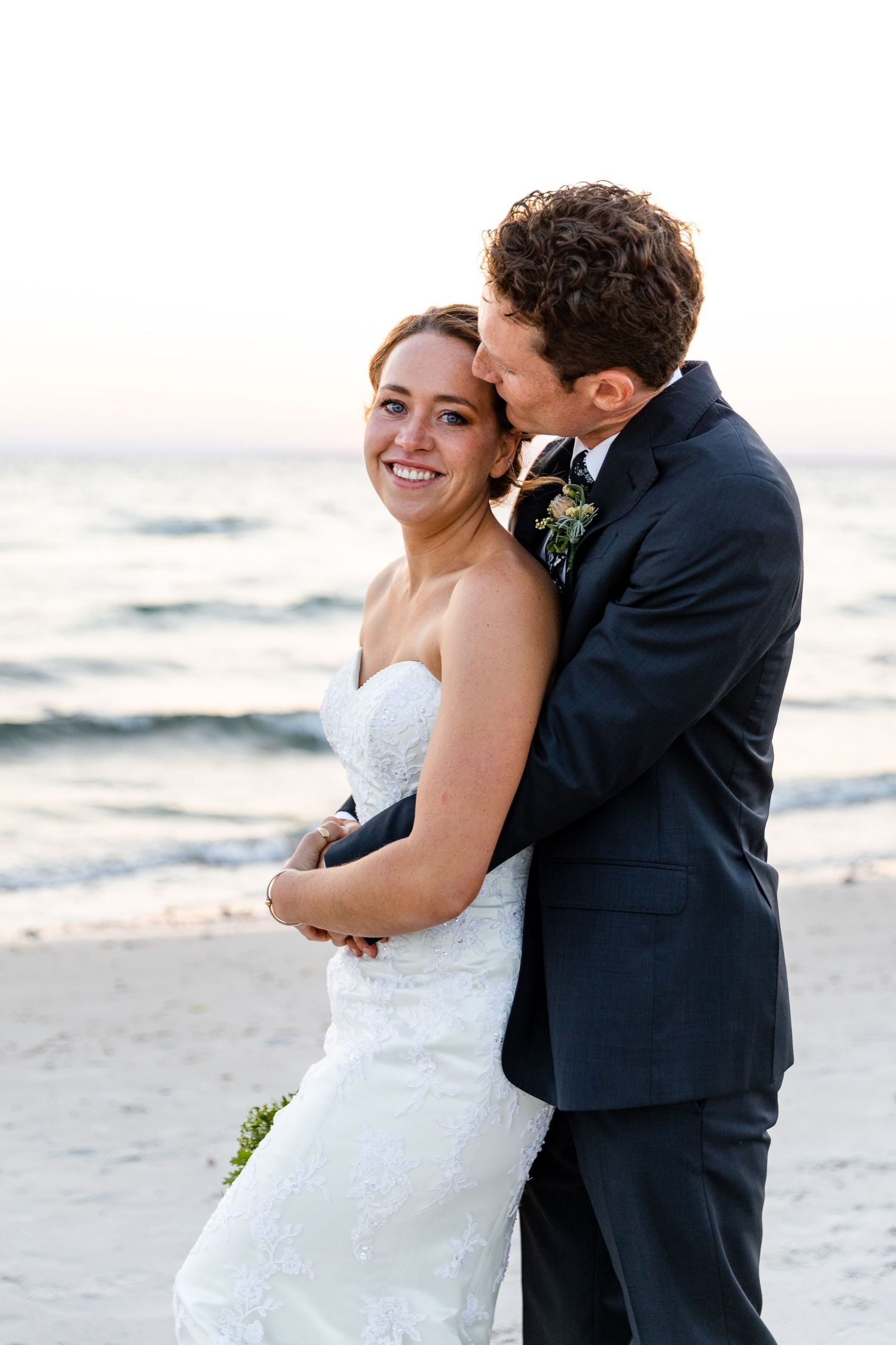 A couple in wedding attire embrace on a beach during sunset as waves roll in along the shore.