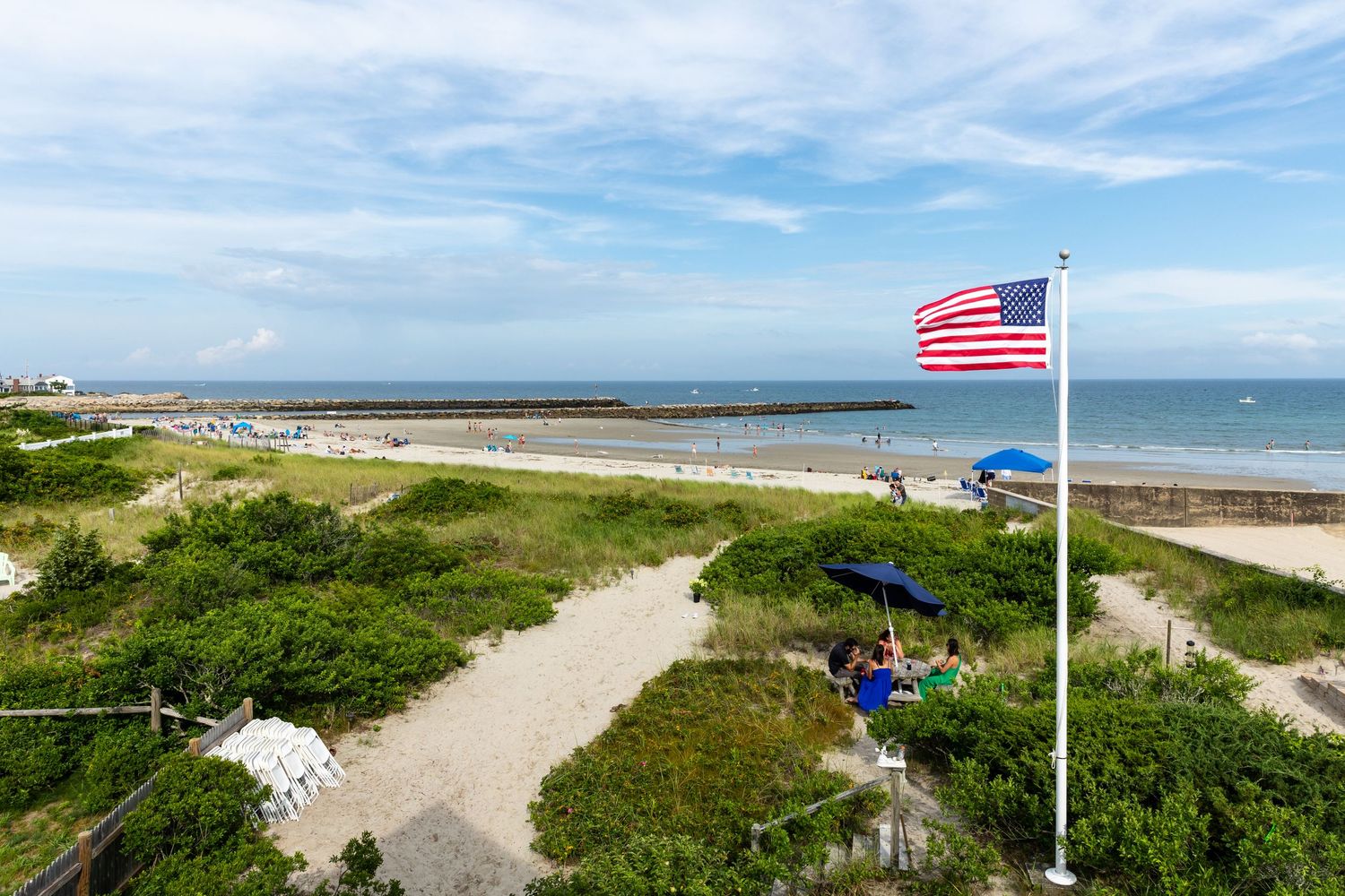 American flag waves over sandy beach path leading to Cape Cod shoreline on sunny summer day with blue skies.