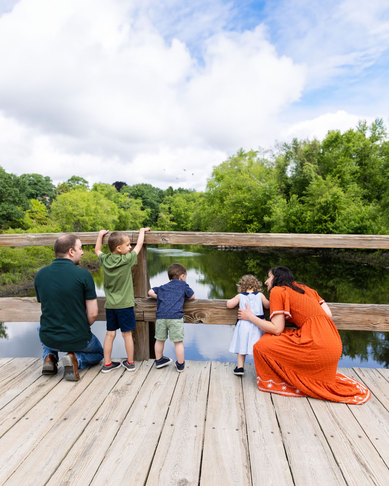 A group of people and children look over a wooden railing at a pond on a sunny summer day.