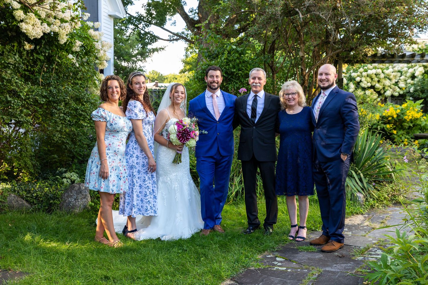 A wedding party poses outdoors on a sunny day surrounded by greenery and garden scenery.