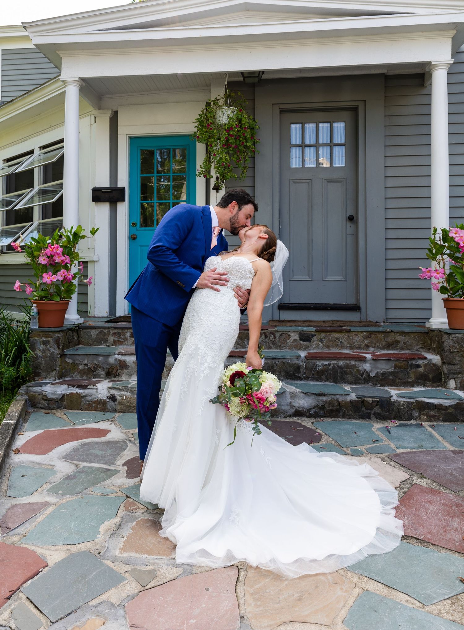 A couple shares an intimate moment on stone steps of a charming gray house with turquoise door and pink flowers.