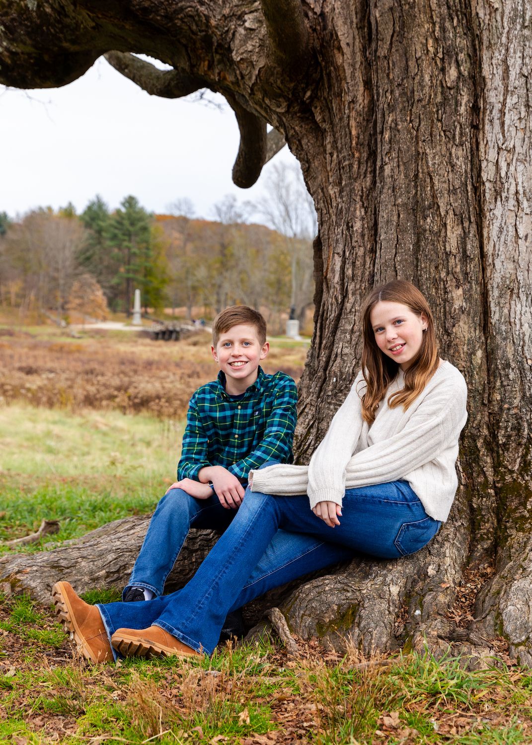 Two young people sit by a large tree trunk in a rural autumn landscape.