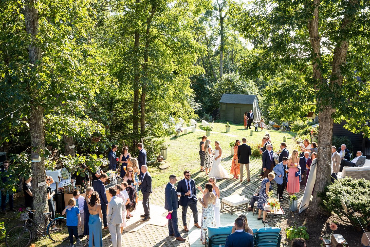 Wedding guests gather outdoors on a sunny garden path surrounded by lush green trees at an elegant summer celebration.