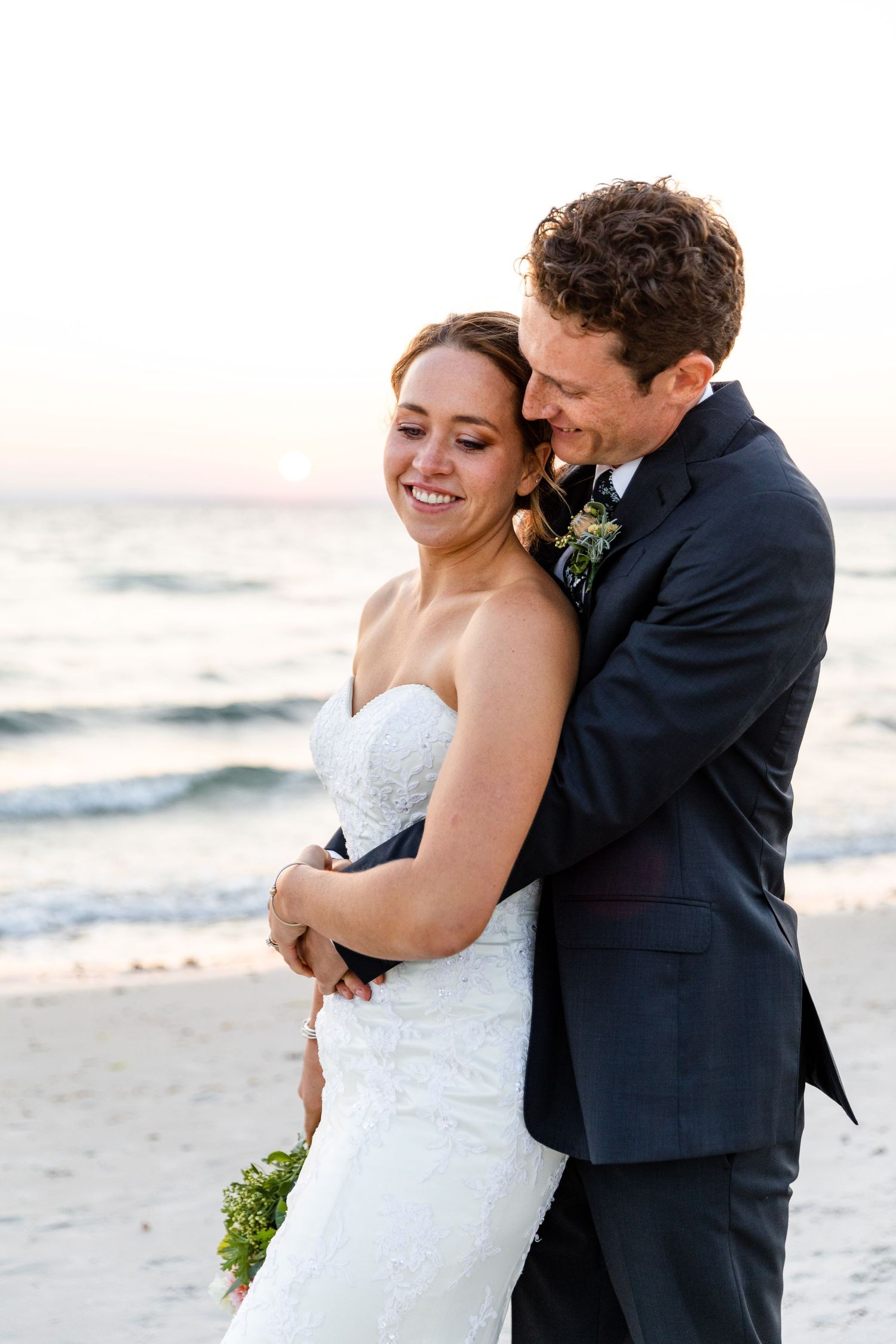 A joyful wedding couple embraces on a beach at sunset in formal attire with ocean waves in the background.
