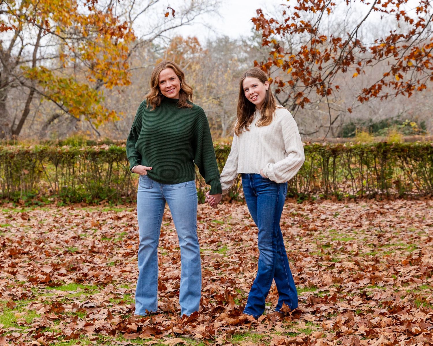 Two people stand together smiling outdoors in a fall setting with autumn leaves on the ground and colorful foliage.