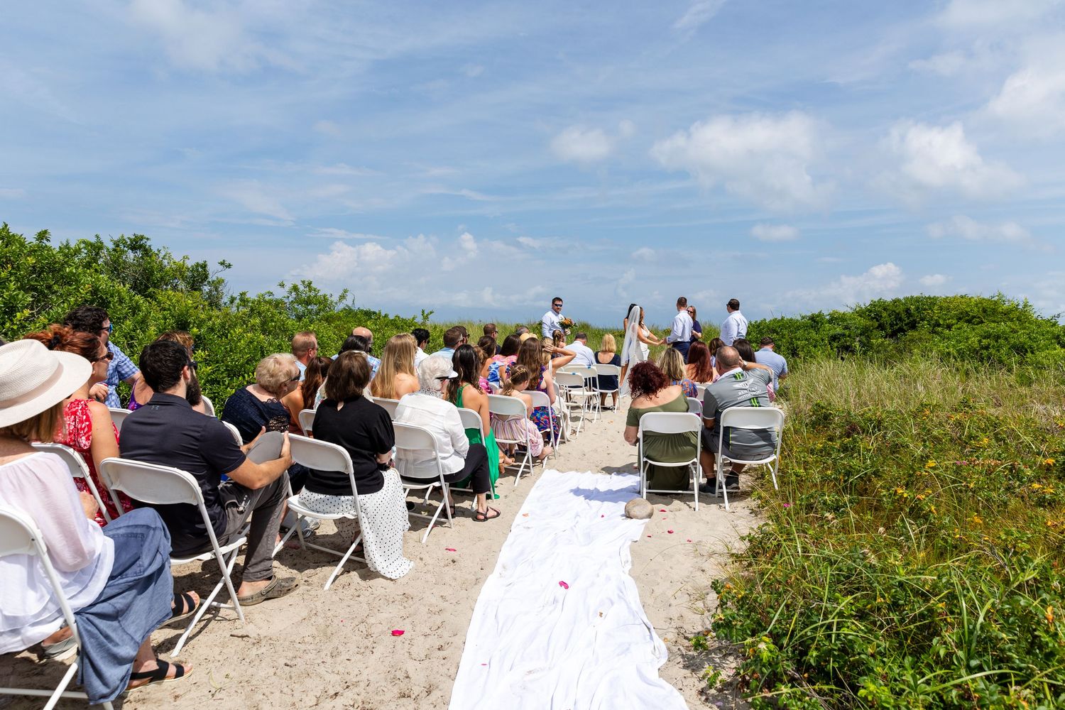 Outdoor beach wedding ceremony with white chairs lined up on a sandy path surrounded by green vegetation on a sunny day.