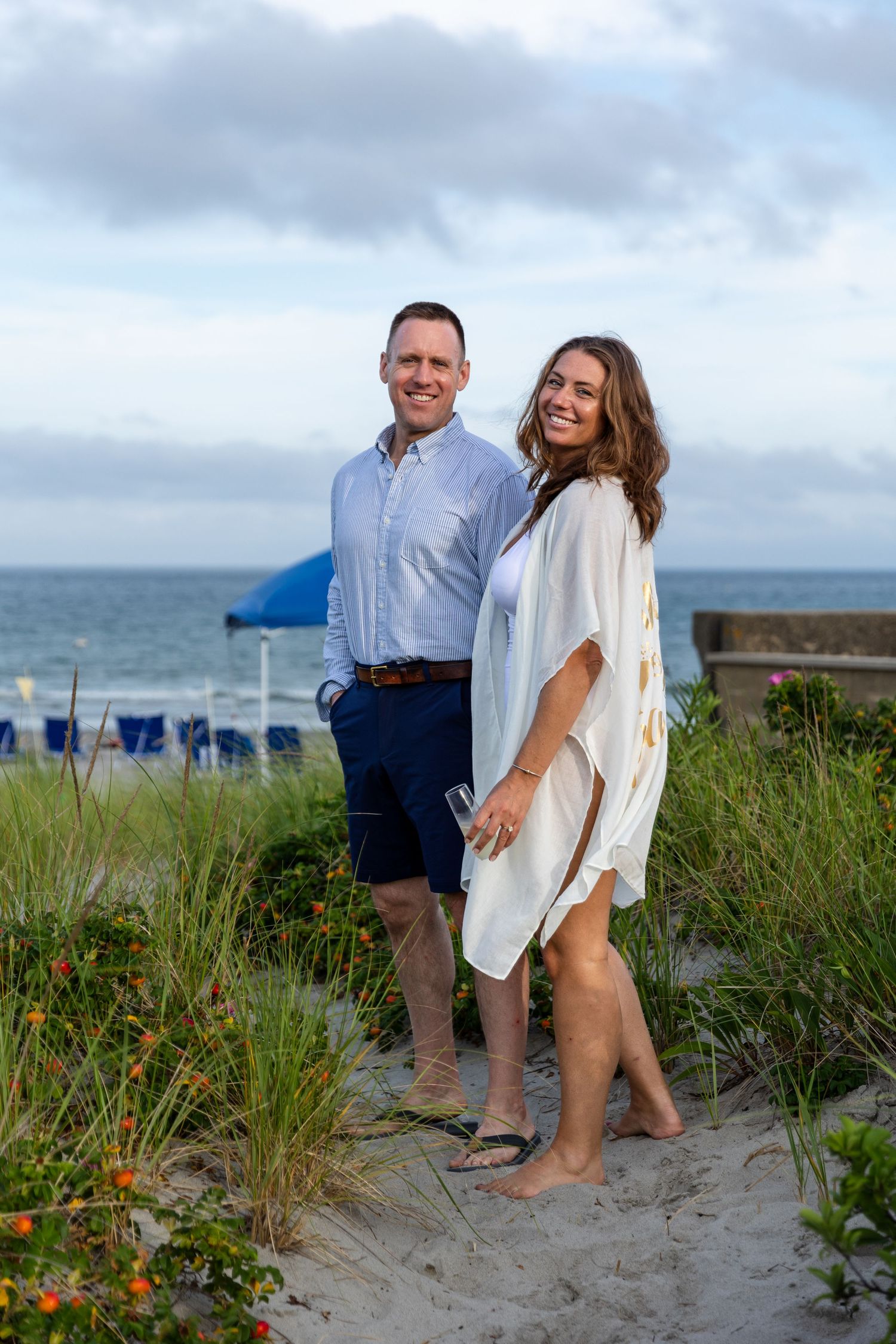 Couple enjoying a casual evening stroll along a sandy beach path with ocean view and cloudy sky in the background.