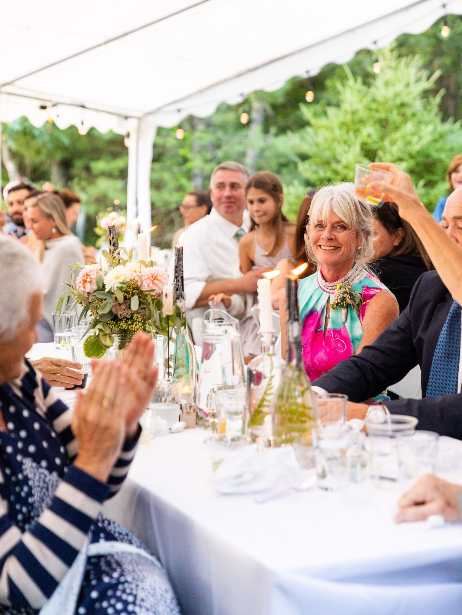 Guests celebrate at an outdoor wedding reception under a white tent with table decorations and string lights.