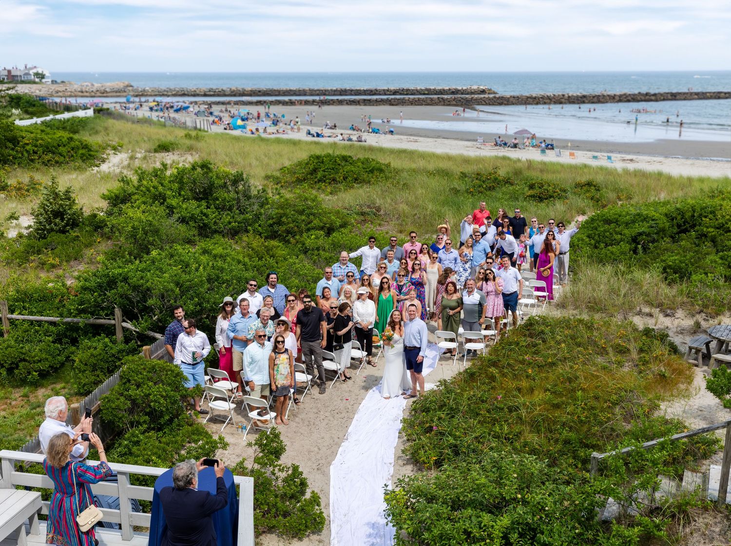 Scenic boardwalk path along the beach with coastal vegetation and groups of beachgoers enjoying a sunny summer day.