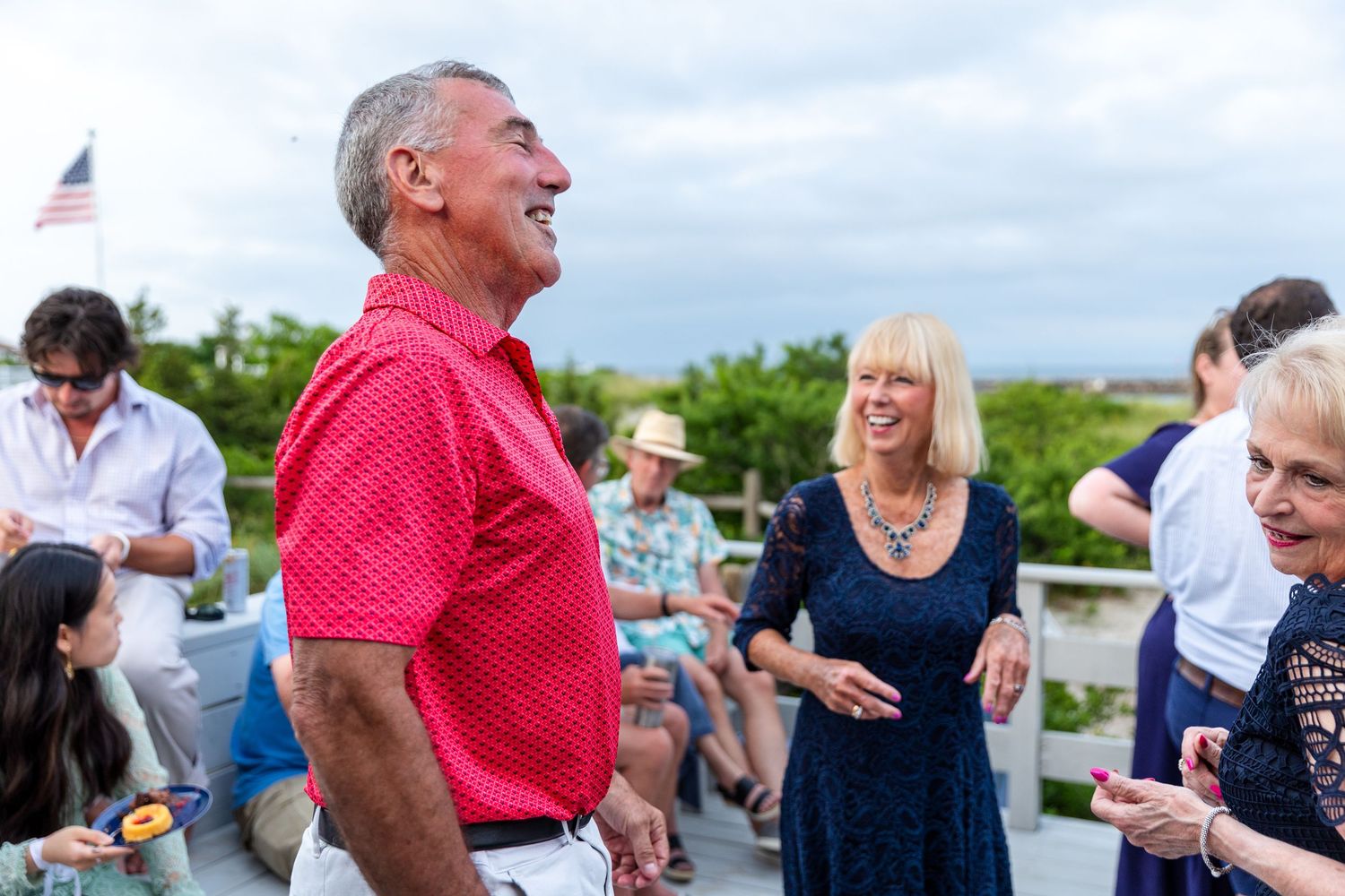 A group of people socialize and laugh together at an outdoor gathering with a flag visible in the background.