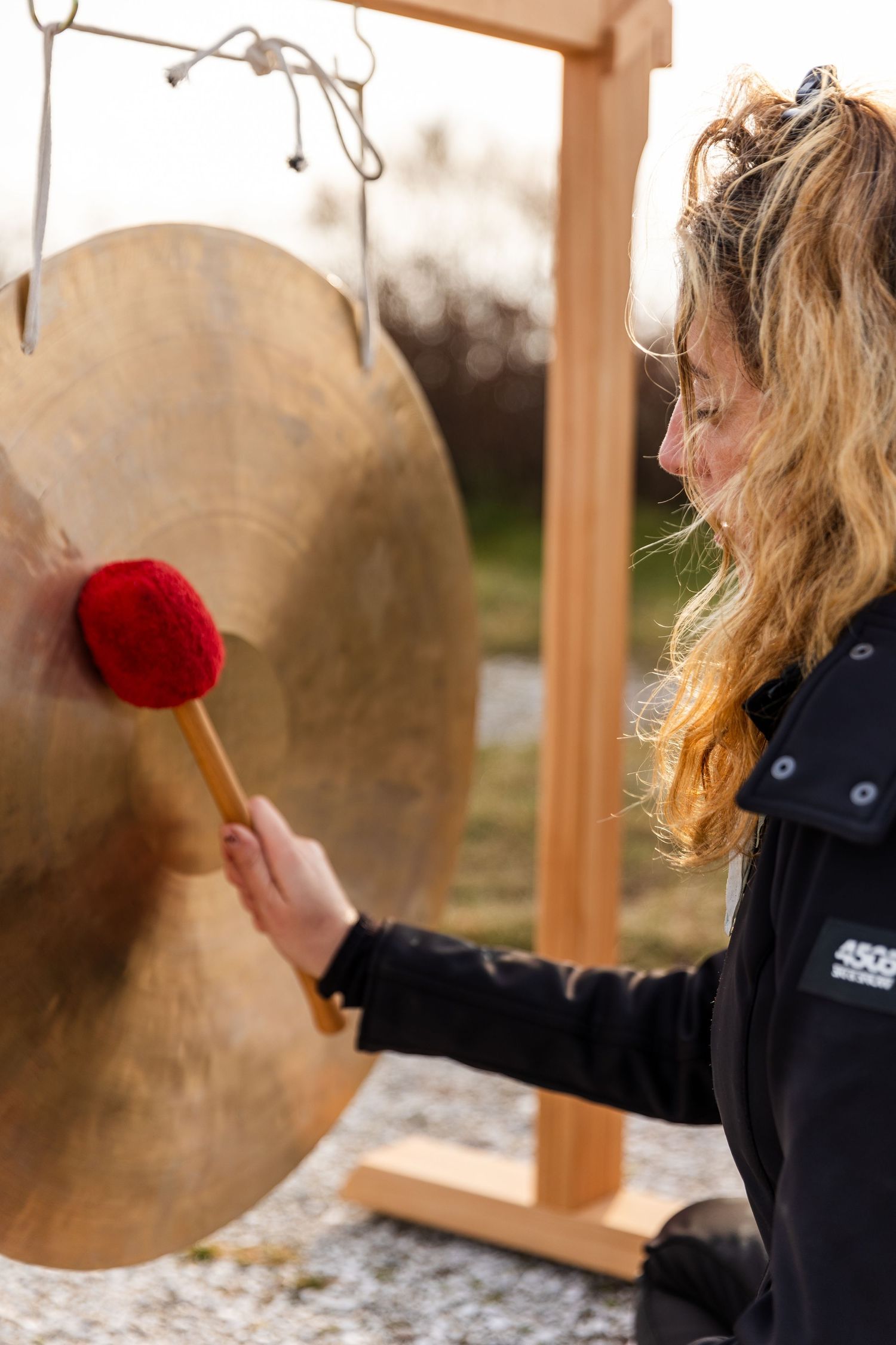 Woman in black jacket uses mallet to strike a large brass gong in an outdoor setting during sunset.