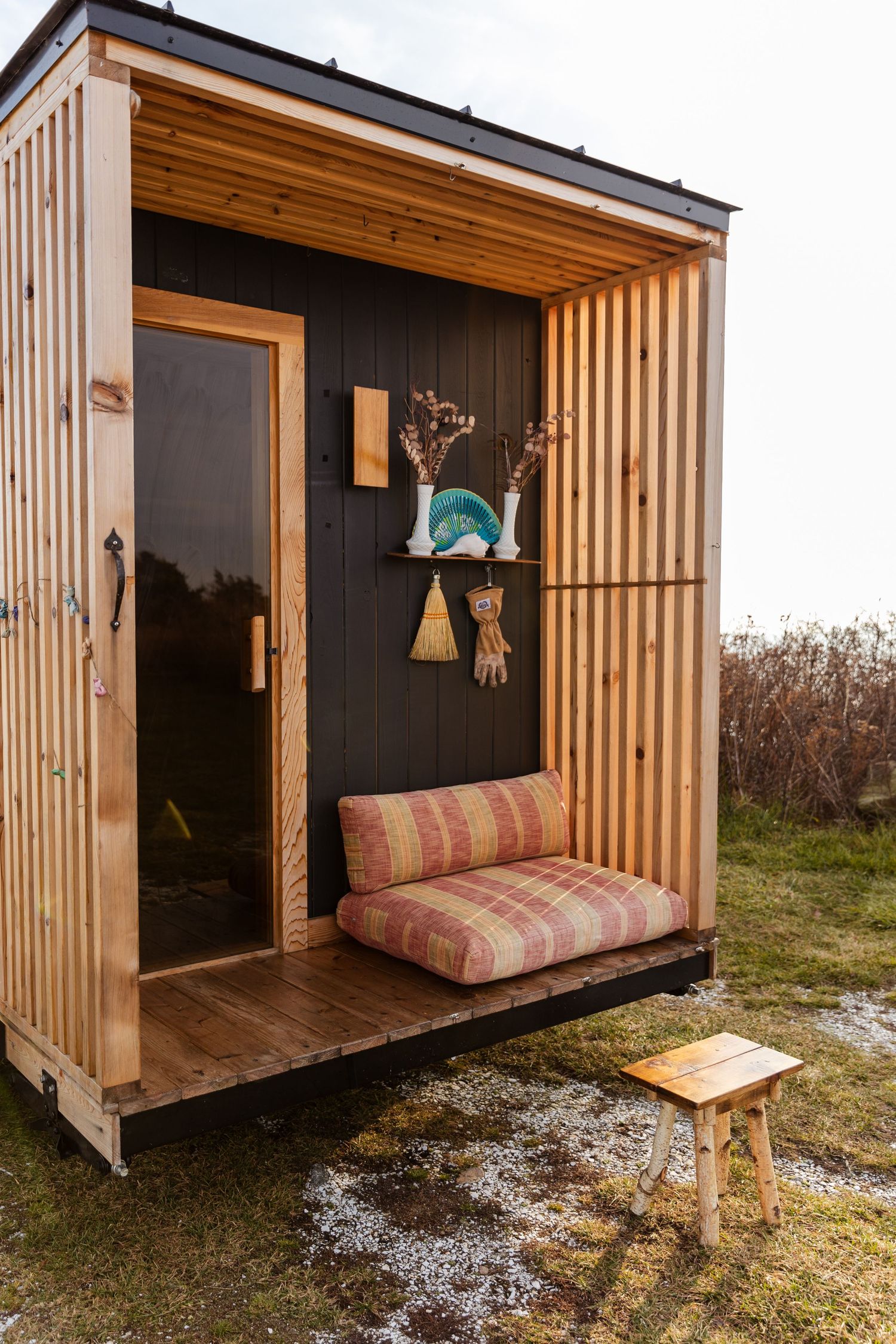Minimalist wooden outdoor shower structure with striped cushion seating and decorative wall hangings in natural setting.