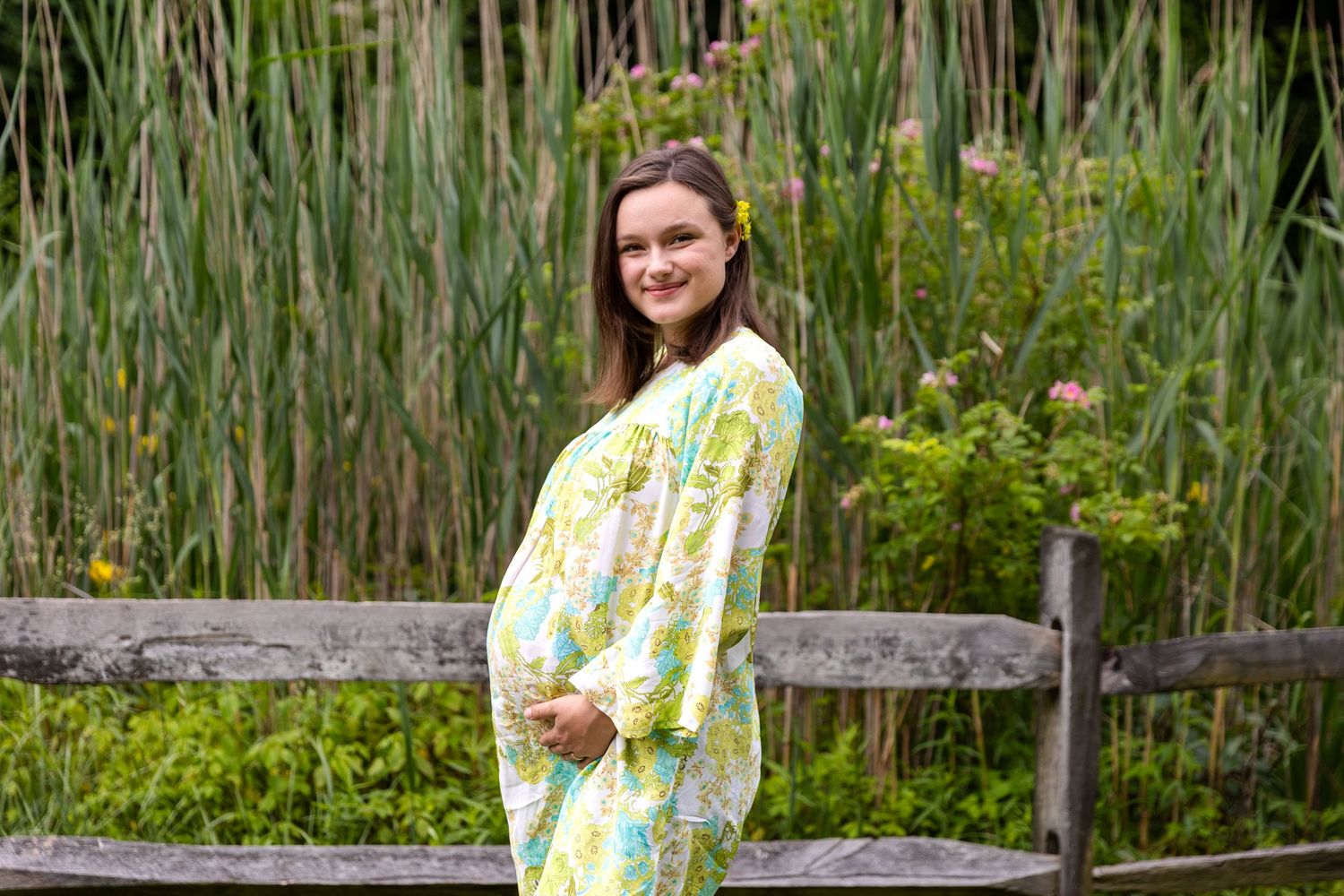 A person wearing a yellow floral dress walks through a lush garden path with tall reeds and wooden fencing.