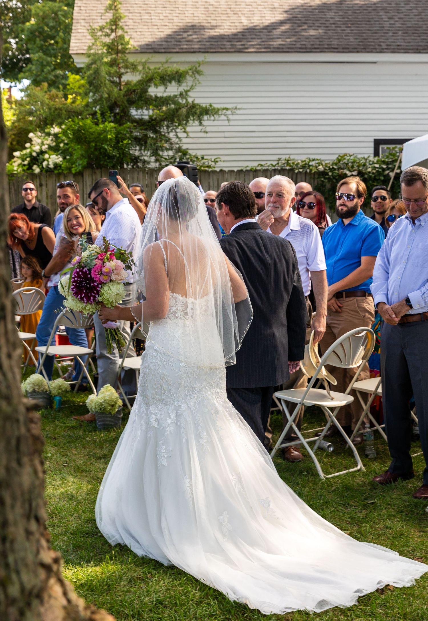A bride in a lace wedding dress with long veil walks on grass during an outdoor backyard ceremony.