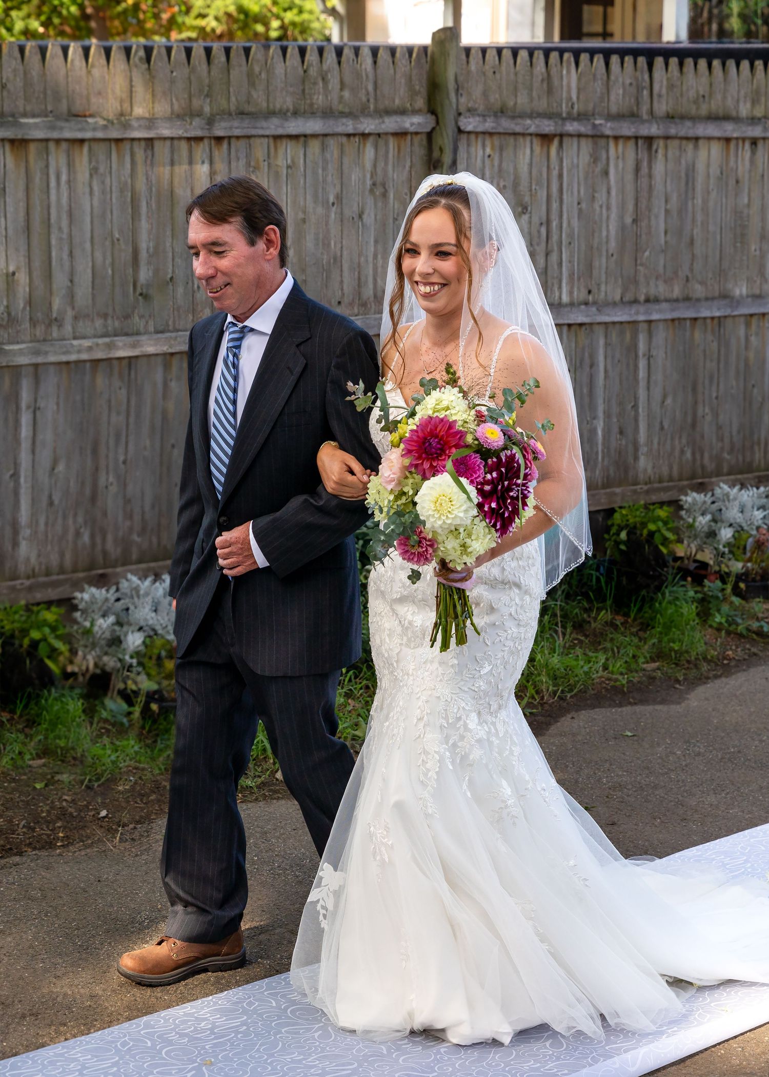 Wedding couple stands in front of a wooden fence while the bride adjusts her dress before the ceremony.