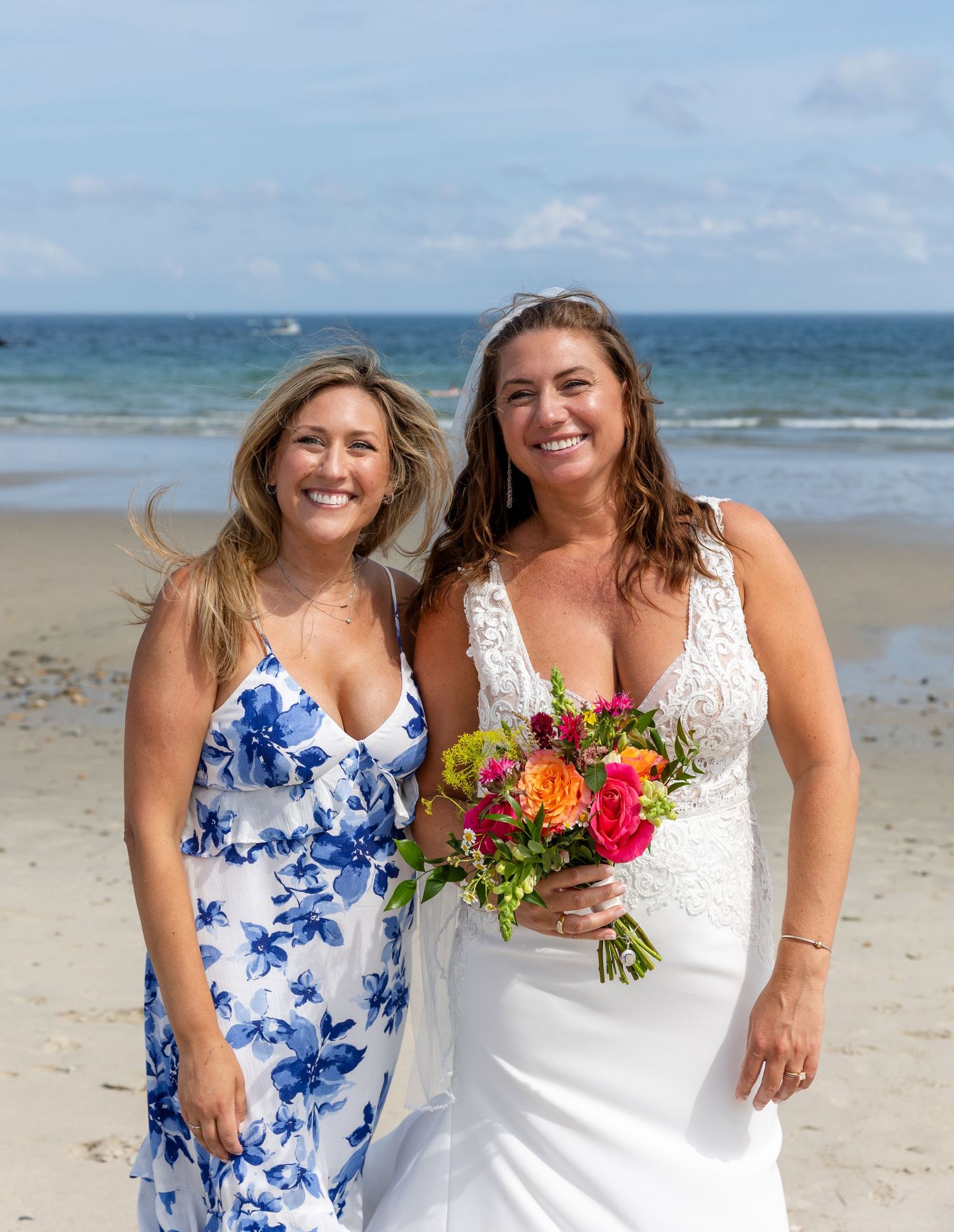 Two wedding guests stand together on a sandy beach with vibrant tropical flowers and ocean in the background.