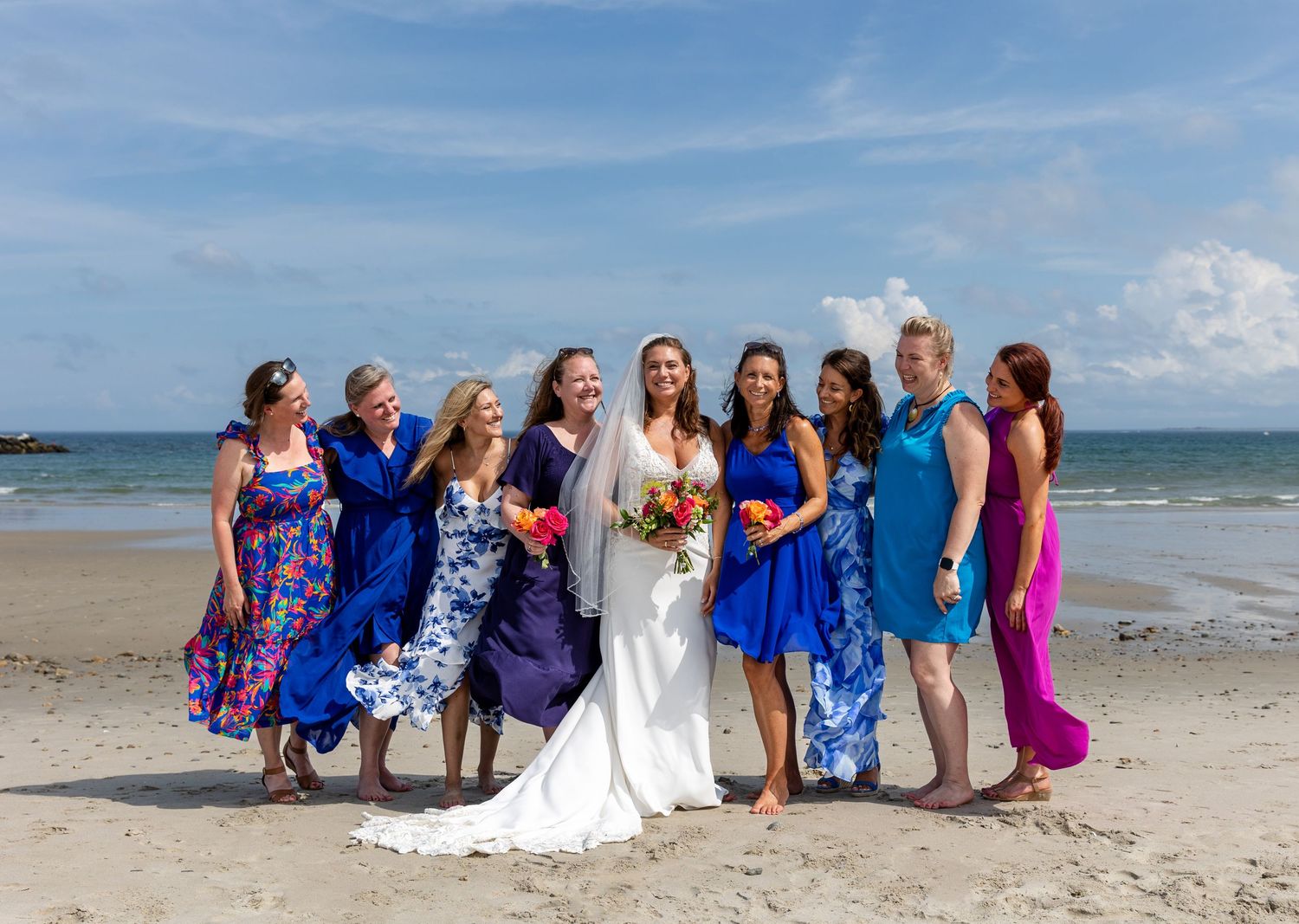 A wedding party stands together on a sunny beach, wearing coordinating blue and purple dresses against an ocean backdrop.