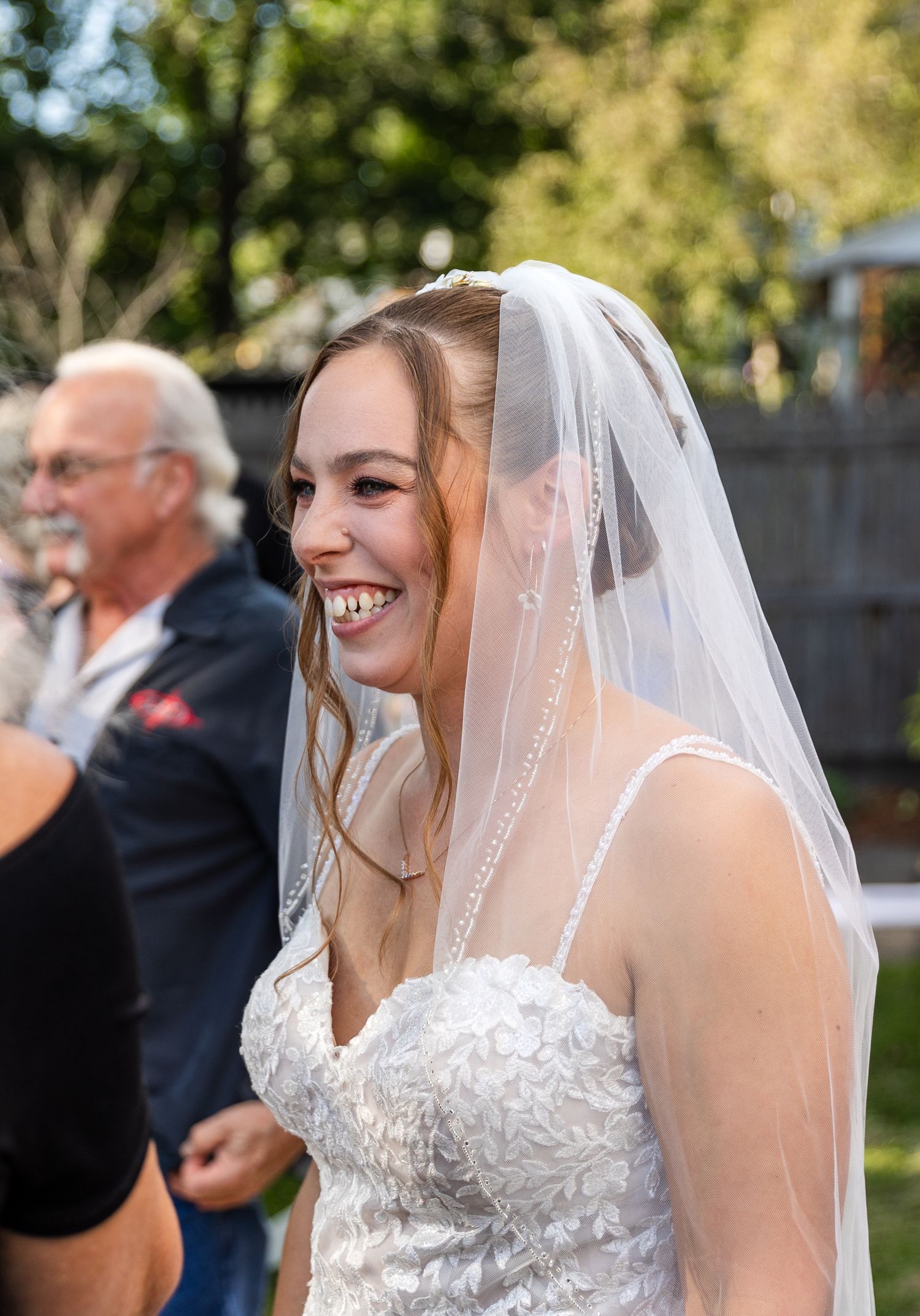 A bride smiles joyfully while wearing a lace wedding dress and white veil during an outdoor garden ceremony.