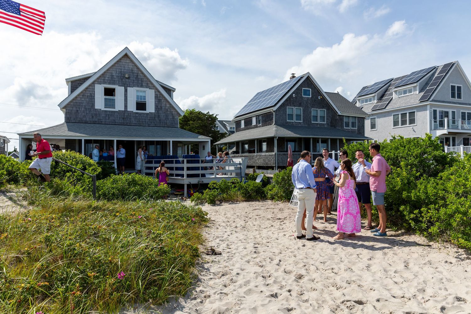 People gather near coastal beach homes on a sandy path with an American flag flying under blue summer skies.