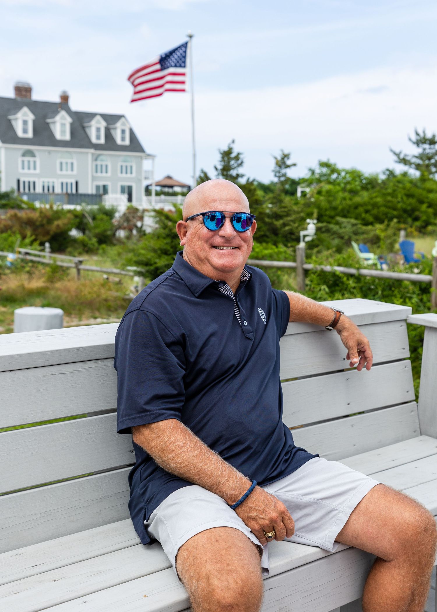 A person in sunglasses and navy shirt reclines on a white bench at a coastal location with American flags visible.