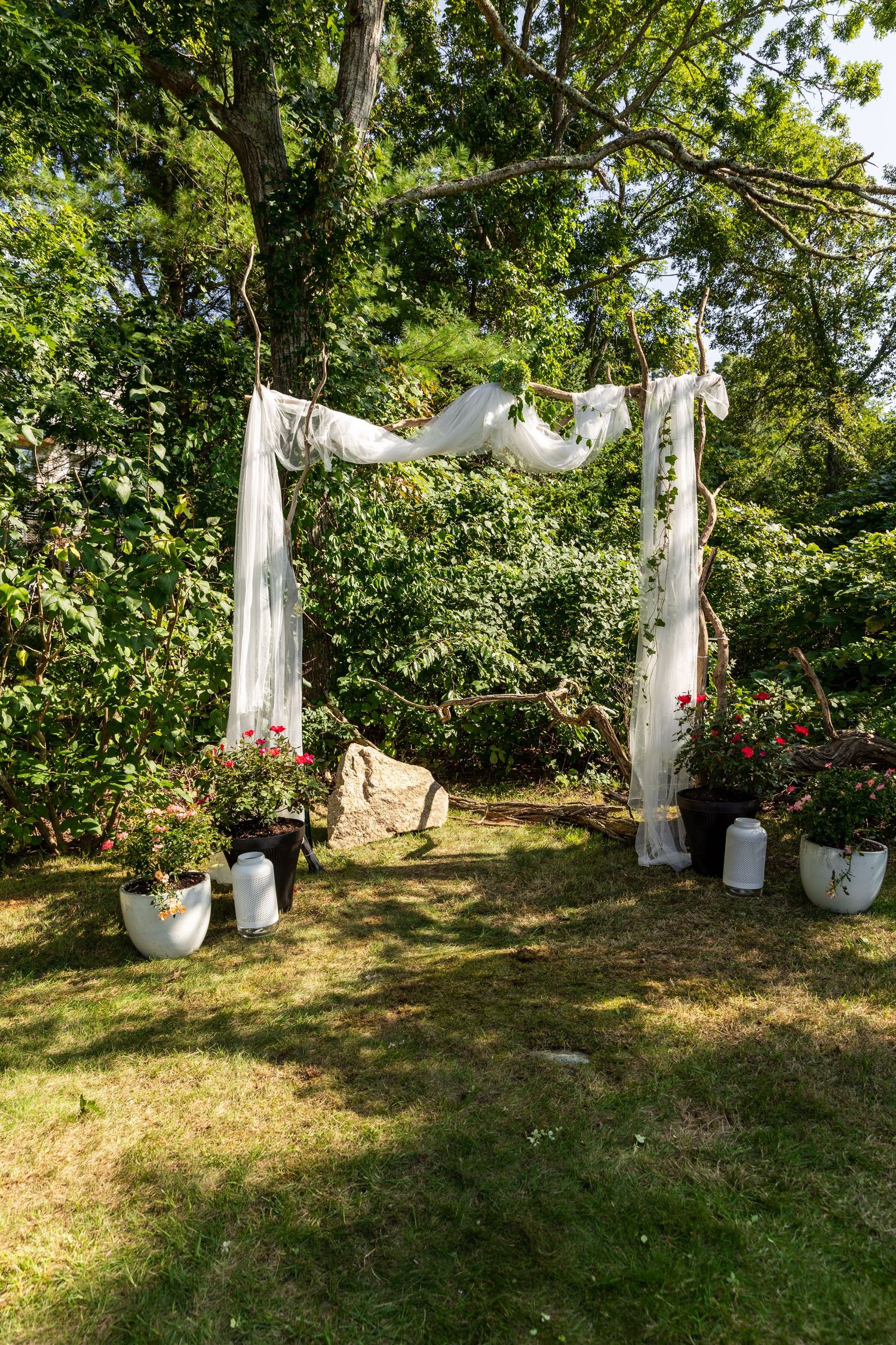 A rustic wedding arch with white fabric draping and flower pots decorating an outdoor garden ceremony space.